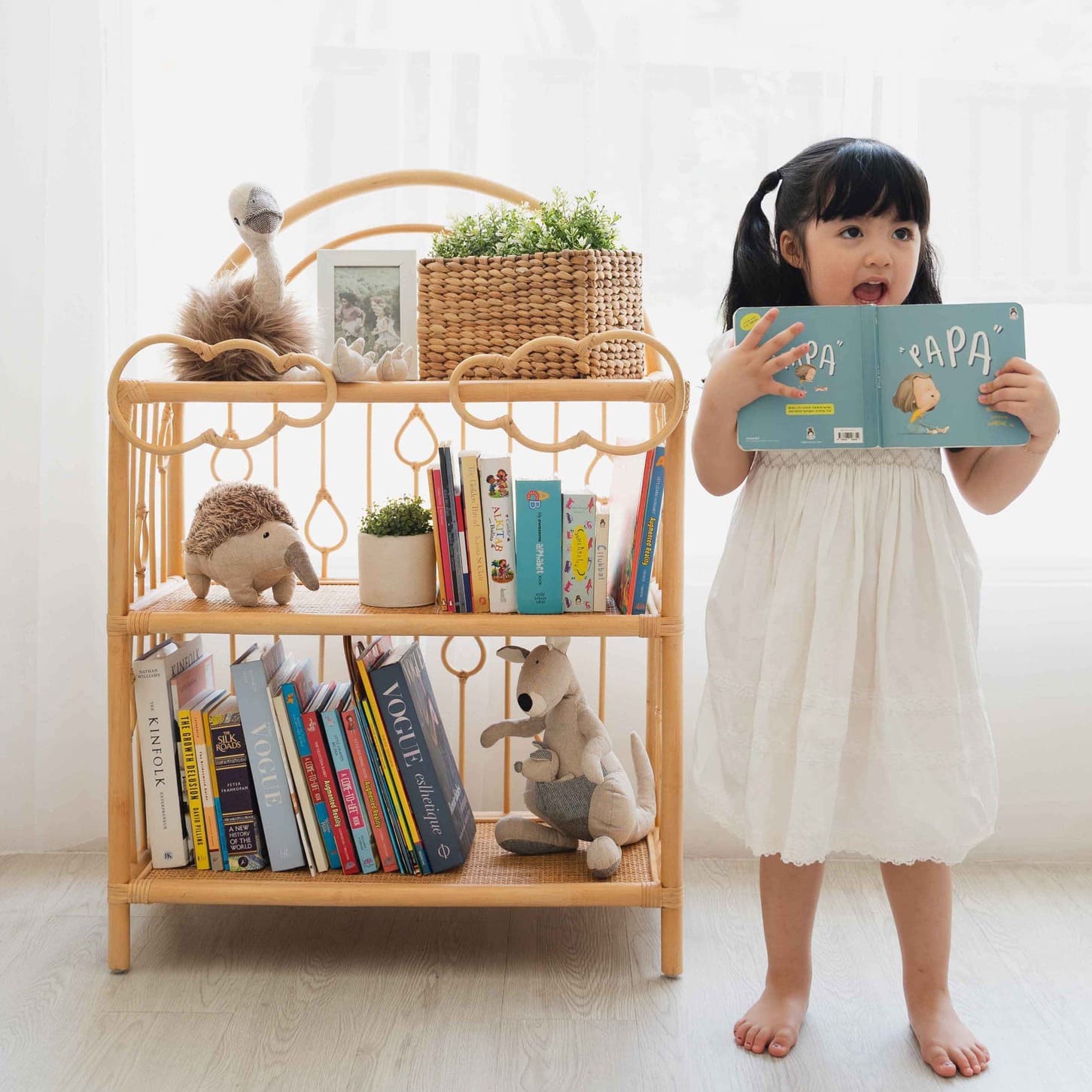 Child holding a book beside the After the Rain Shelf by MOMIJI filled with books and toys indoors.