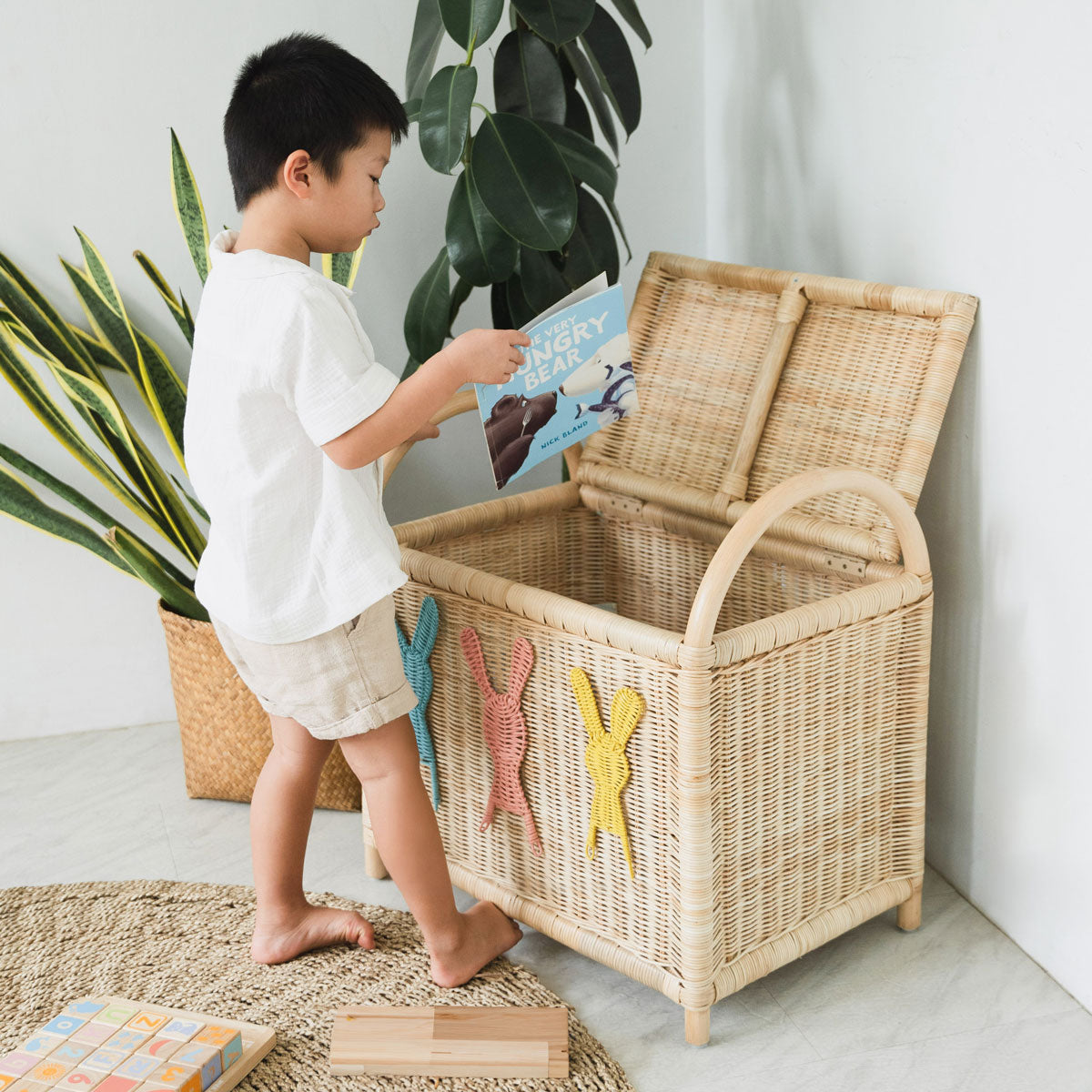 Child packing books and toys into a rattan wicker toy chest in a playroom