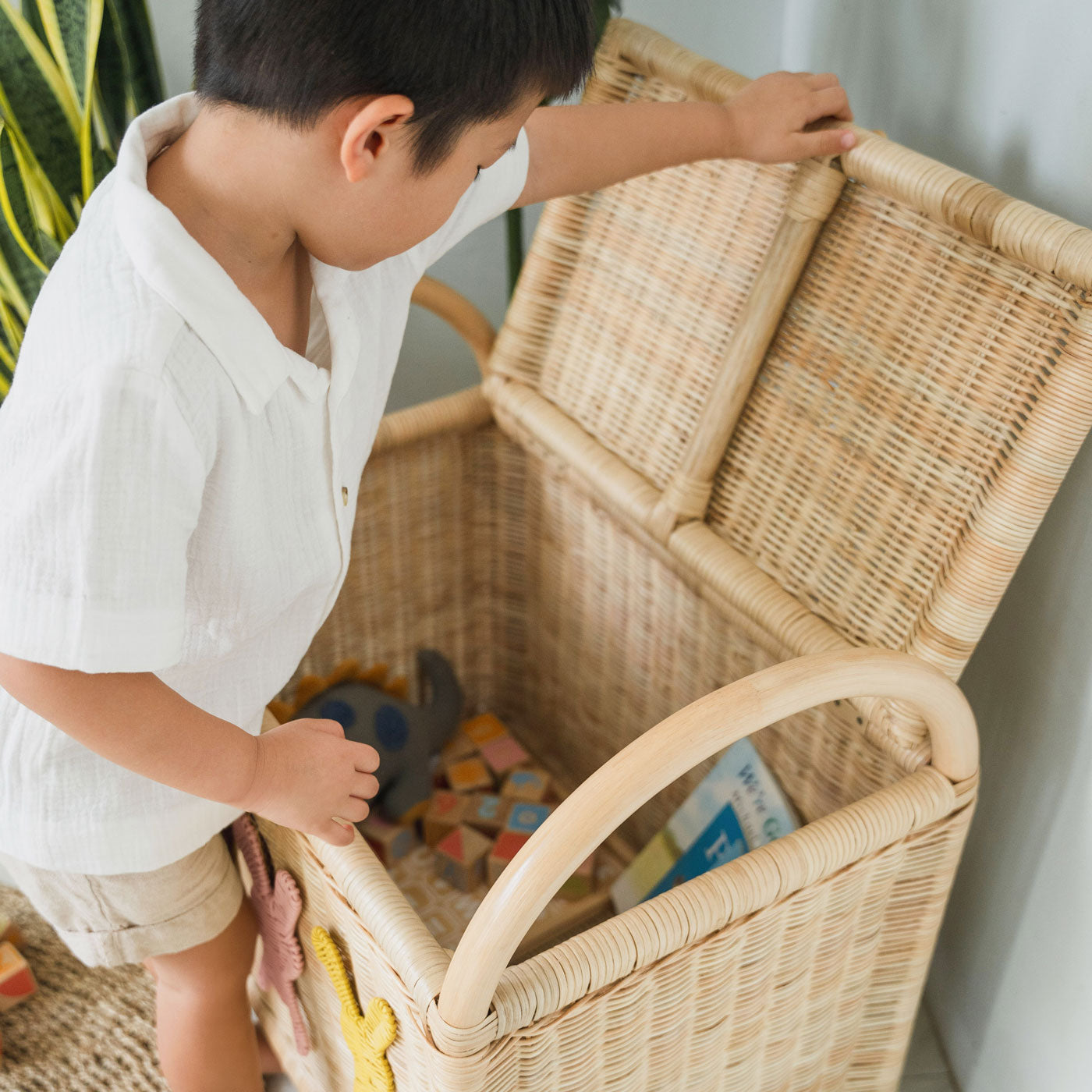 Child opening the lid of a rattan wicker trunk filled with soft toys and wooden blocks