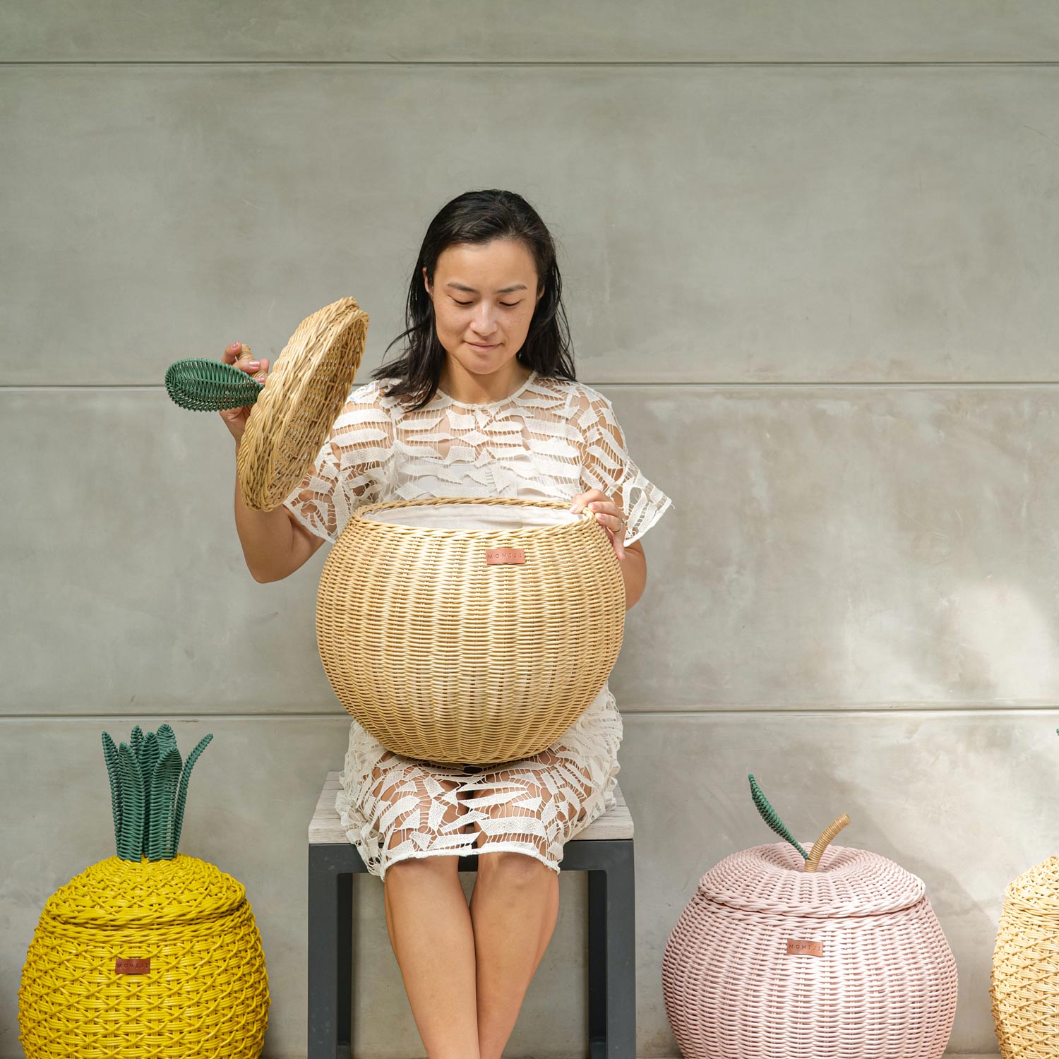 Woman opening a handcrafted natural rattan apple storage basket in medium size by MOMIJI, styled with yellow pineapple and pink apple baskets on the floor.