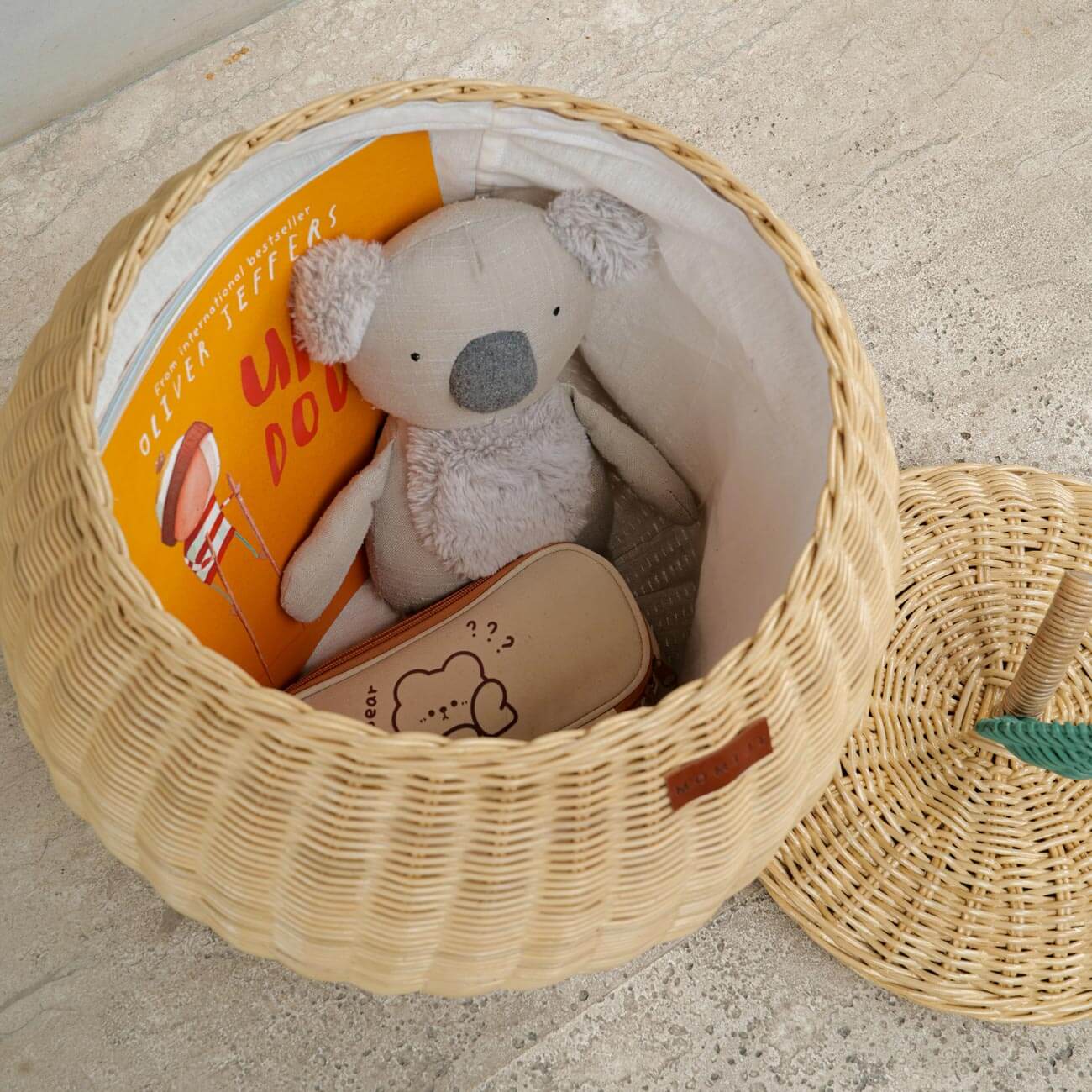 Top view of a handcrafted natural rattan apple storage basket by MOMIJI with the lid off, filled with a book and plush toy, showing the white inner fabric lining on a concrete surface.