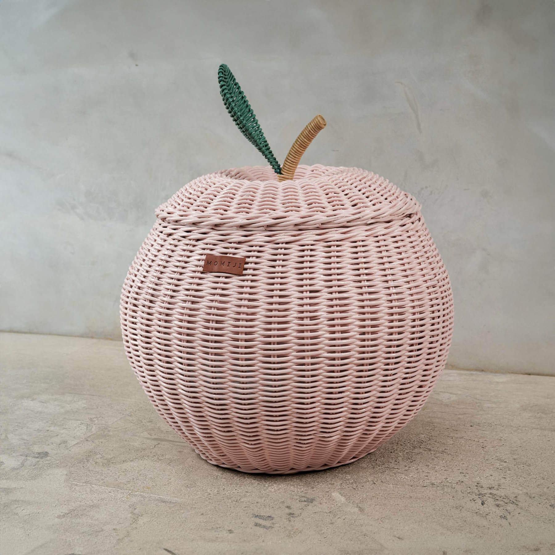 Titled view of a handcrafted pink rattan apple storage basket in medium size by MOMIJI, highlighting the woven design, MOMIJI label, and lid with green leaf and natural stem.