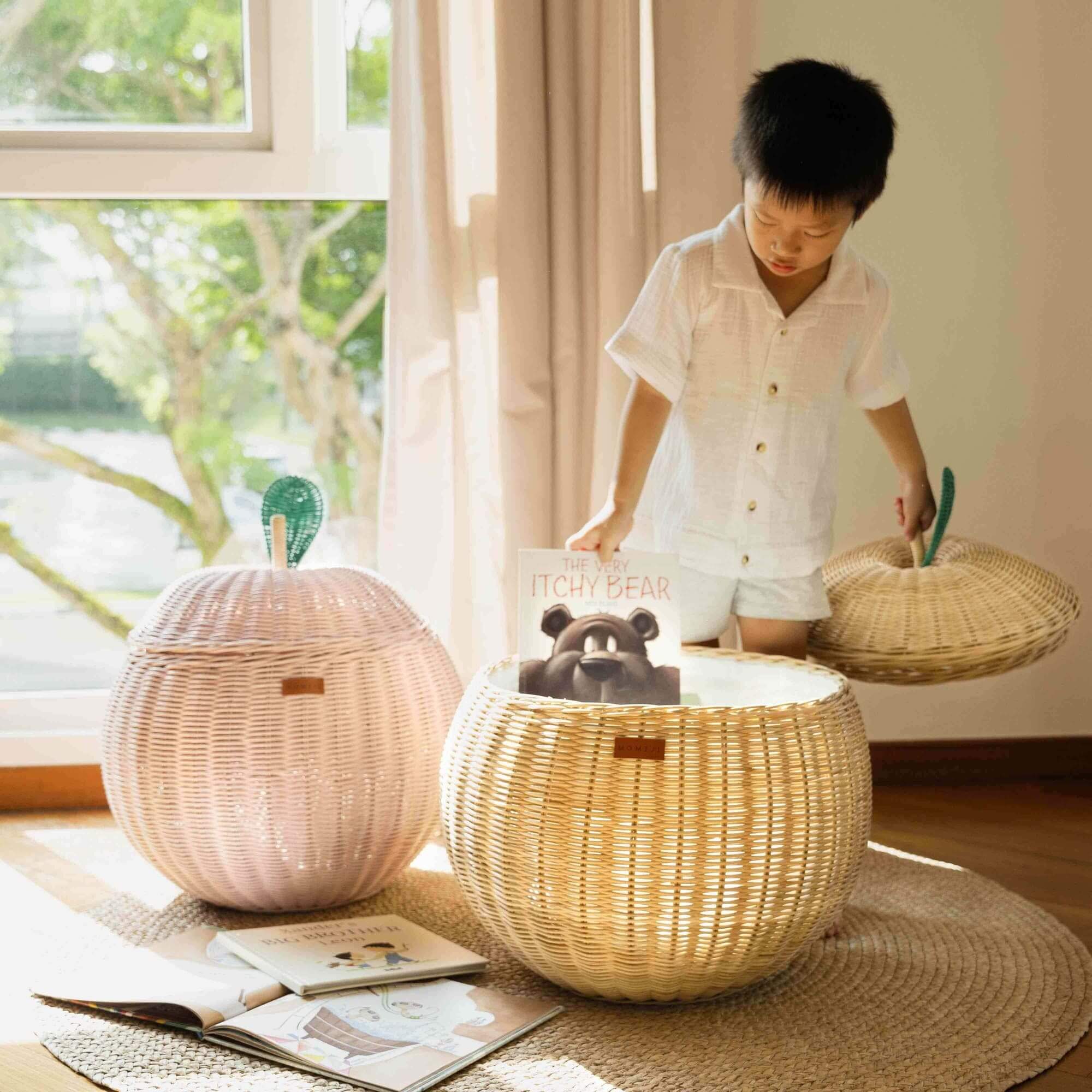 Child opening the natural apple rattan storage basket while holding the lid and picking up a book inside the rattan basket beside the pink apple rattan storage basket by MOMIJI.