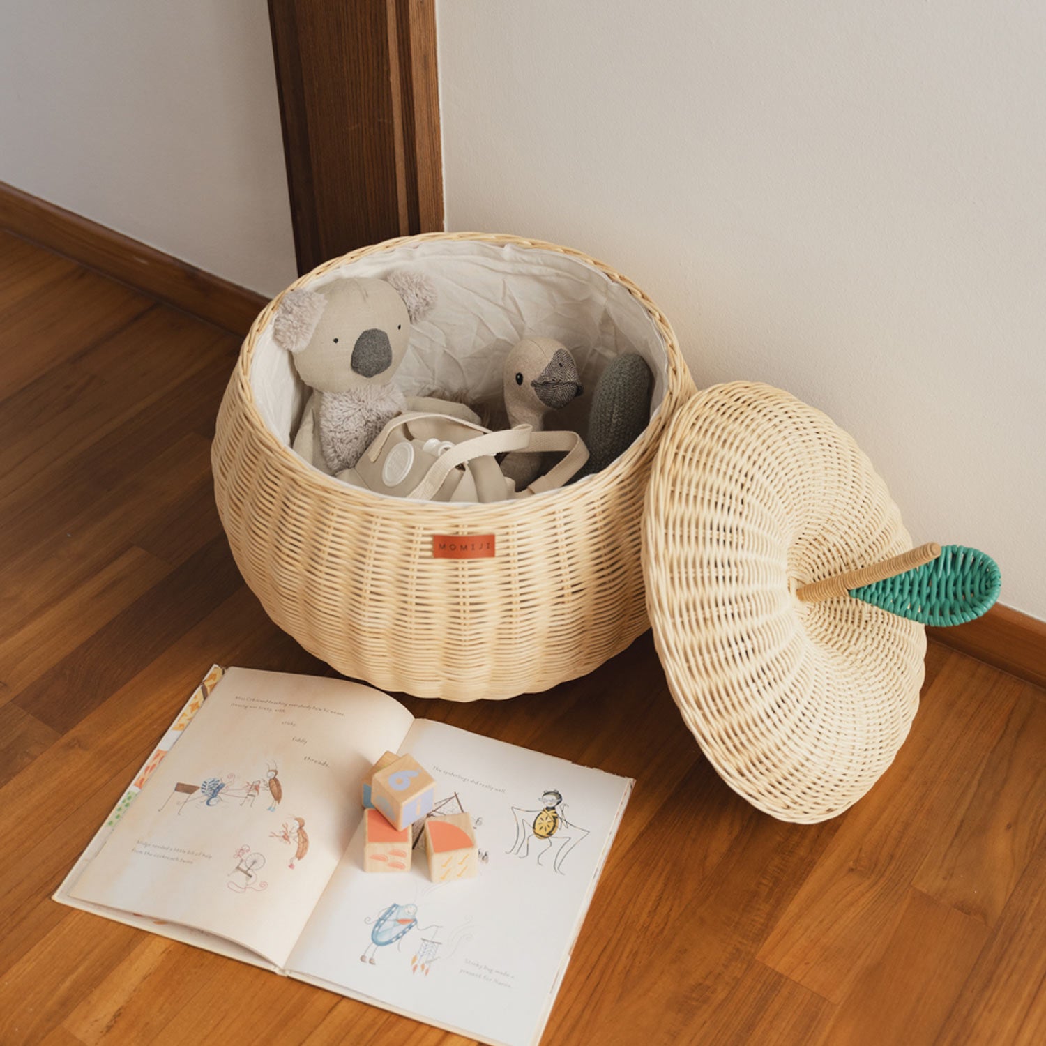 Top view of an opened natural apple rattan storage basket by MOMIJI with a lid that has an apple leaf on it with a mini bag and stuffed toys inside, on a wooden floor next to a book and wooden blocks.