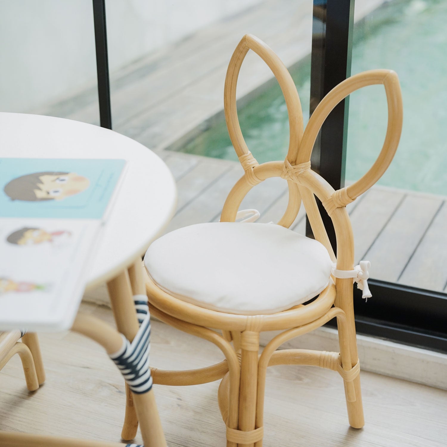 A handcrafted natural rattan Aulia Kids Table paired with a Butterfly Kids Chairs in natural, featuring butterfly-shaped backrests and soft cushions, styled in a bright indoor setting beside large glass doors overlooking an outdoor swimming pool.