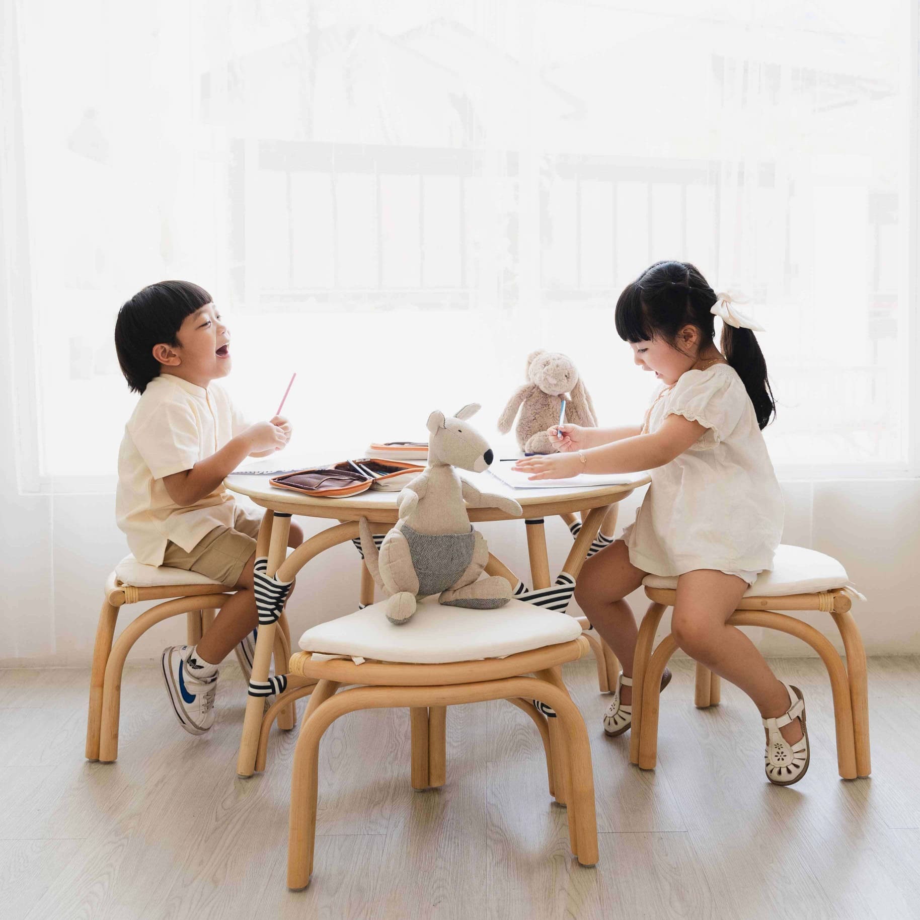 Two children sitting at Aulia Table Set with Stools by MOMIJ with toys and coloring tools in a bright room.