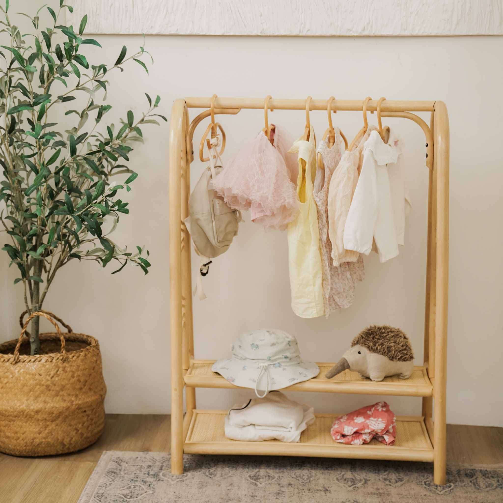 Handcrafted natural rattan Belle Kids Montessori Clothing Rack by MOMIJI featuring two storage shelves and hanging hooks, styled with baby clothes, a hat, a stuffed hedgehog, and folded textiles in a nursery-inspired room.