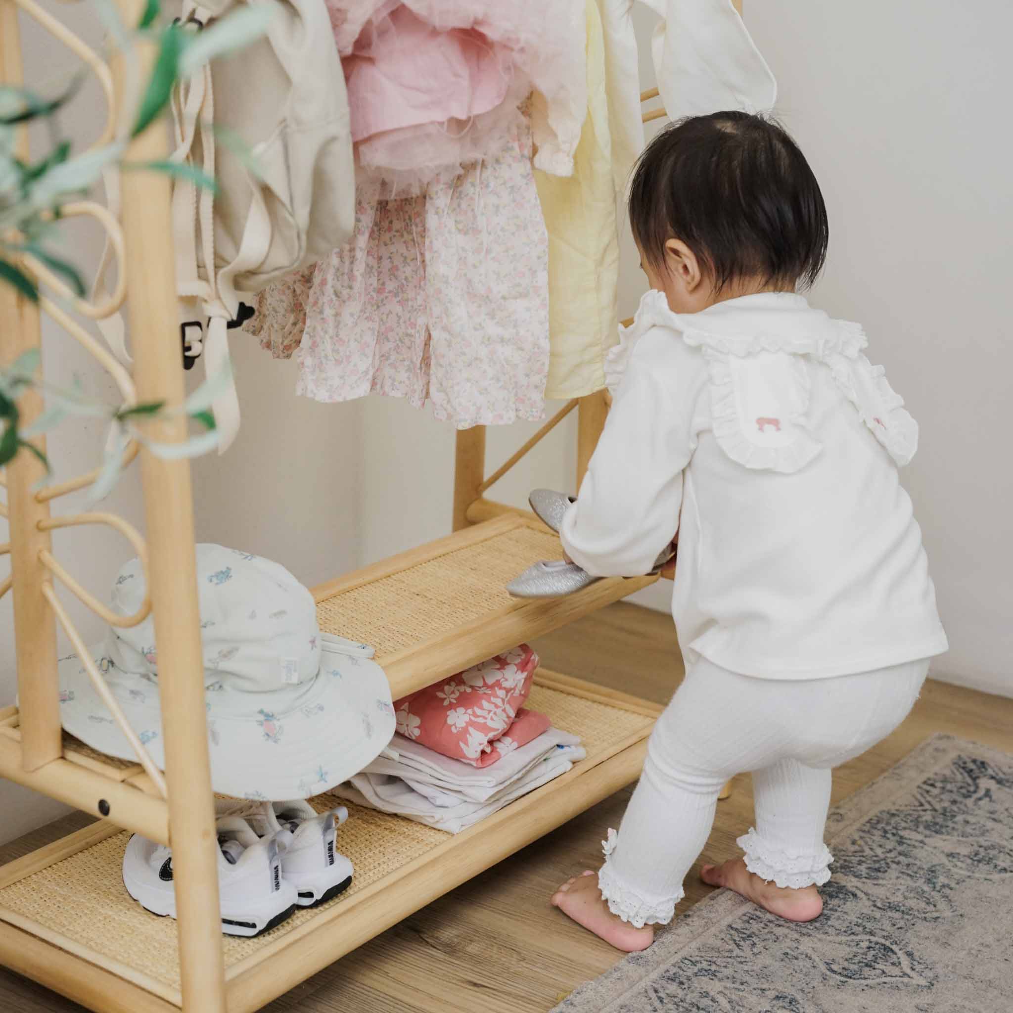 A toddler reaching towards the handcrafted natural rattan Belle Kids Montessori Clothing Rack by MOMIJI, highlighting the two lower shelves designed for easy-access storage of children's shoes and belongings.