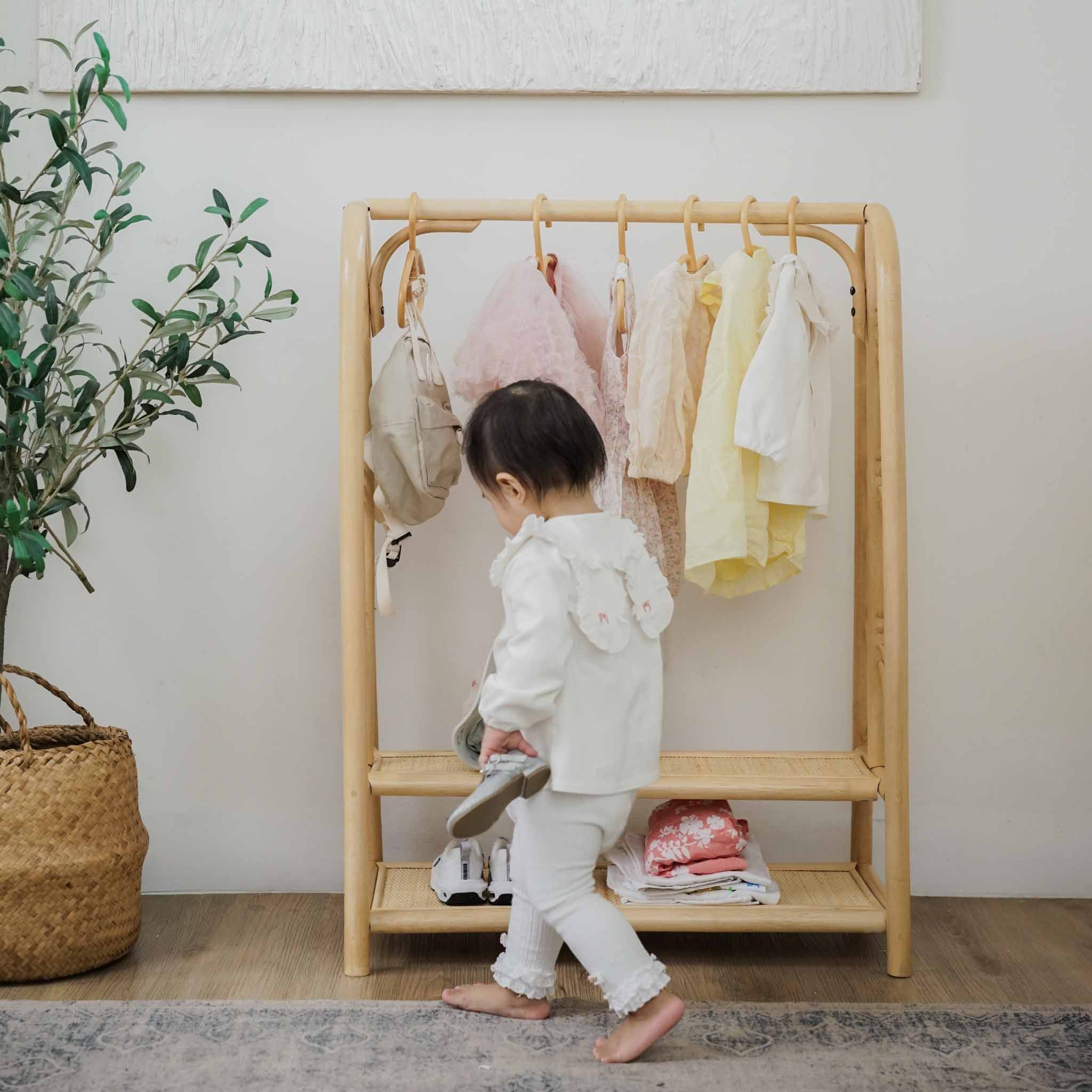 A toddler placing shoes on the shelves of the handcrafted natural rattan Belle Kids Montessori Clothing Rack by MOMIJI, showcasing its multifunctional design for storing children's clothing, shoes, and everyday essentials in a bright room.