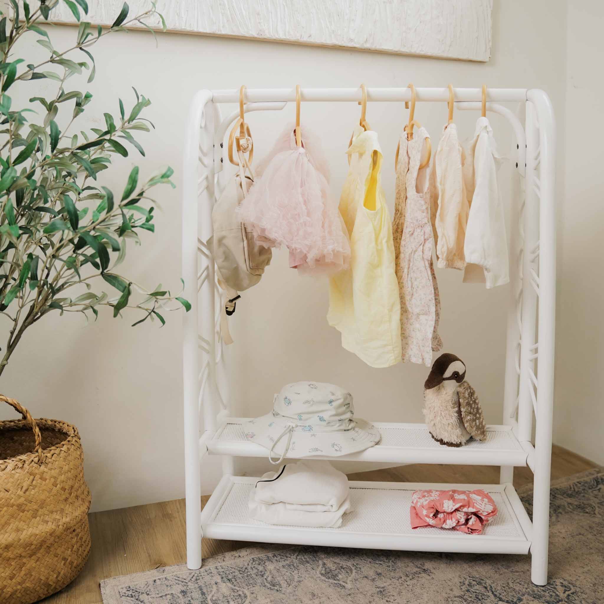 Handcrafted white rattan Belle Kids Montessori Clothing Rack by MOMIJI featuring a hanging rail and two storage shelves, styled with baby garments, a sun hat, a plush toy, and folded textiles in a nursery-inspired room.
