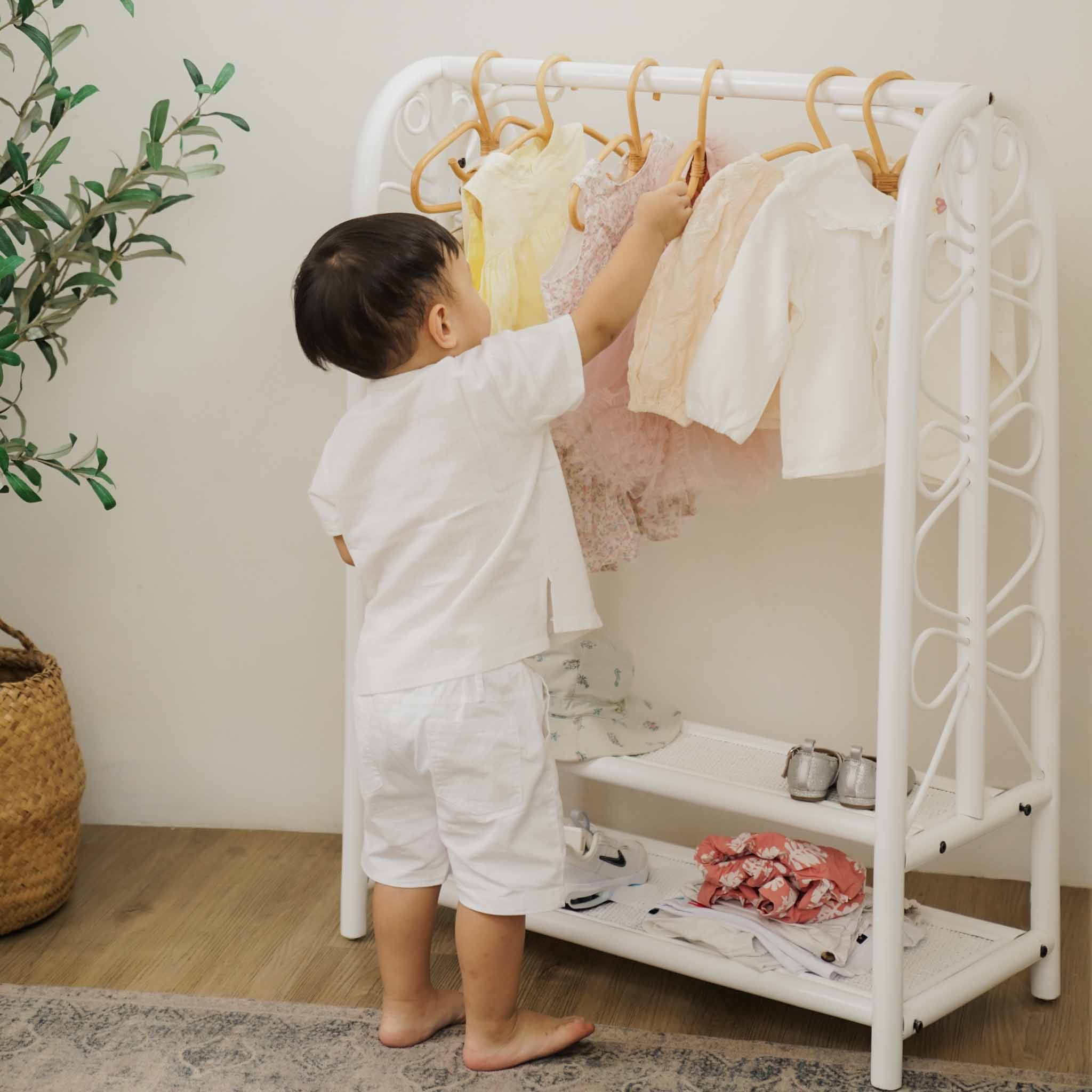 A toddler reaching for clothing on the handcrafted white rattan Belle Kids Montessori Clothing Rack by MOMIJI, highlighting its child-friendly hanging rail and two storage shelves in a bright modern room.
