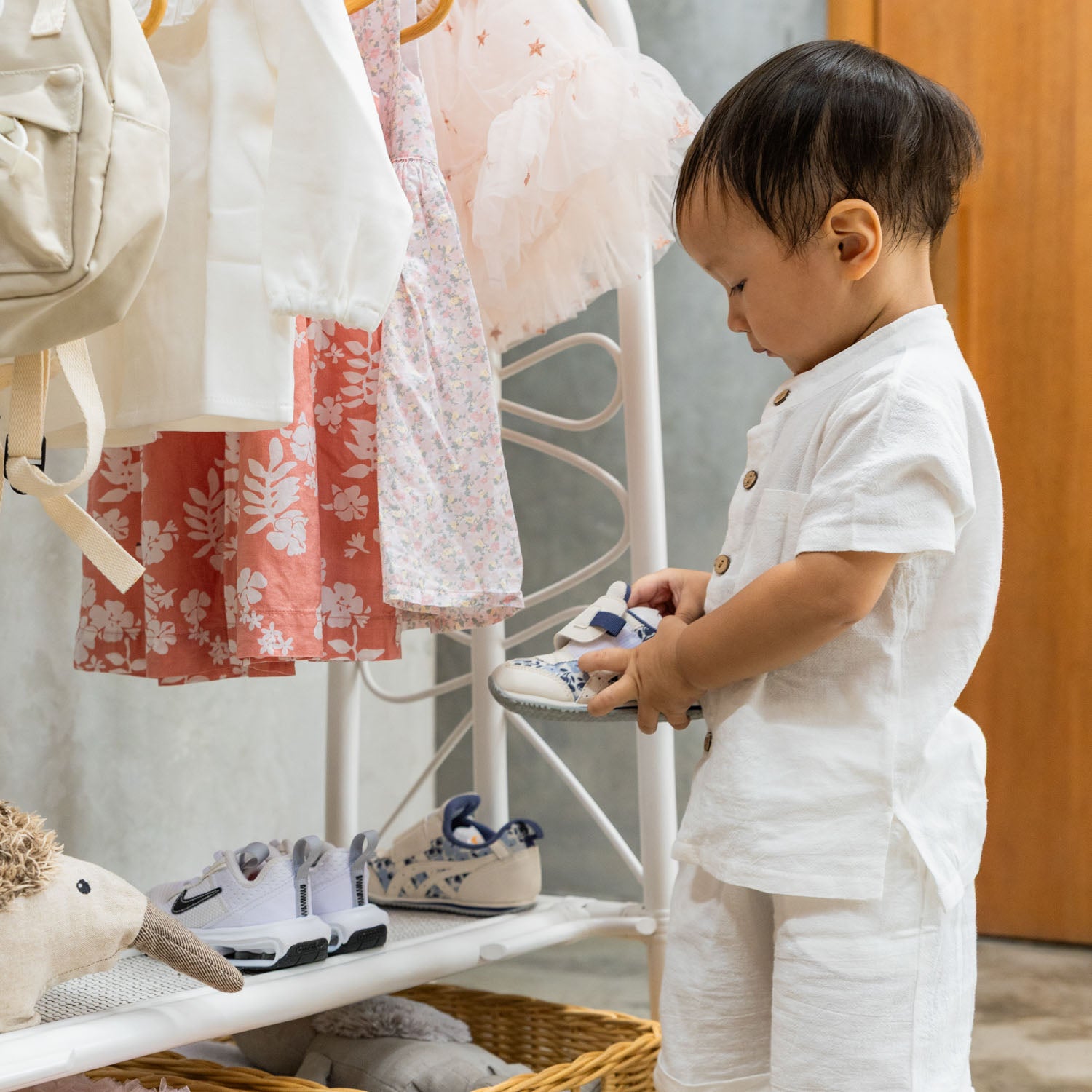 A toddler standing beside the handcrafted white rattan Belle Kids Montessori Clothing Rack by MOMIJI, holding a shoe while additional shoes are neatly stored on the lower shelves, showcasing multifunctional storage for children's essentials.