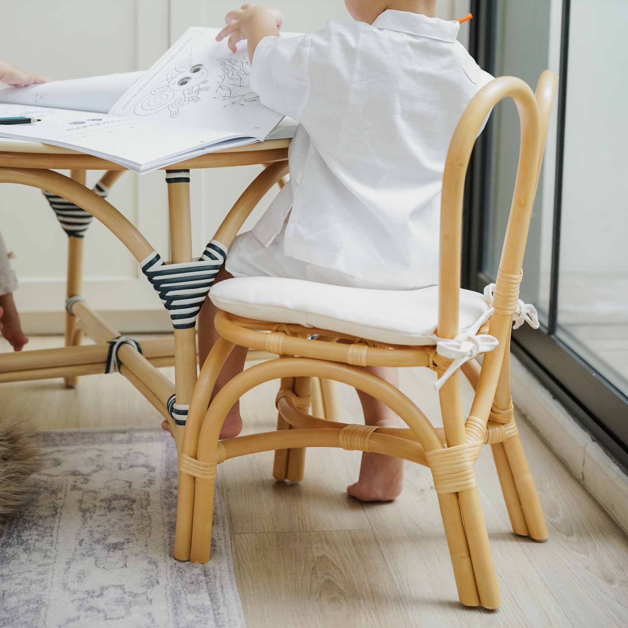 Side view of a child sitting comfortably on a handcrafted natural rattan wicker Bunny Kids Chair with a soft removeable cushion, paired set with the Aulia Kids Table (Medium) by MOMIJI in a Montessori-inspired playroom.