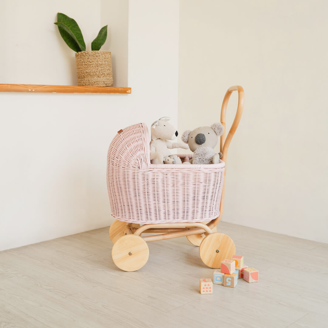 Side view of handcrafted pink rattan wicker Colette Doll Stroller by MOMIJI filled with stuffed animals featured wooden wheels, styled on a wooden floor.