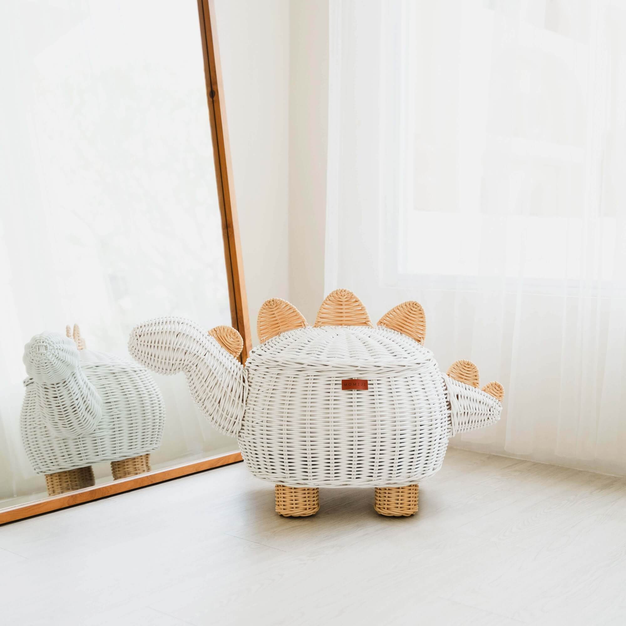 A white dinosaur rattan storage basket by MOMIJI placed on a wooden floor next to a mirror, with a reflection of a similar basket in the mirror.