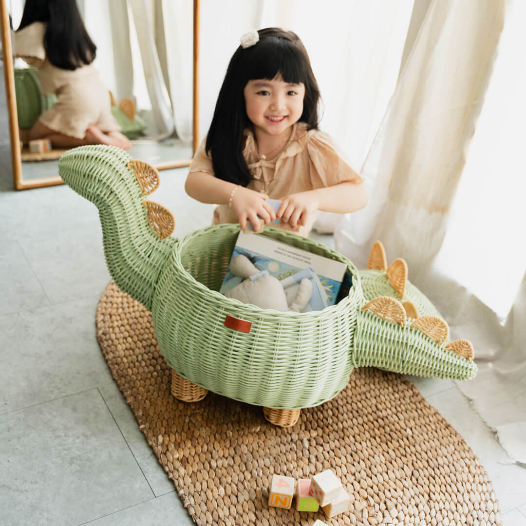 A child playing with the Mint Green Dinosaur Storage Rattan Basket by MOMIJI with books and toys inside the rattan basket and a mirror behind the child.