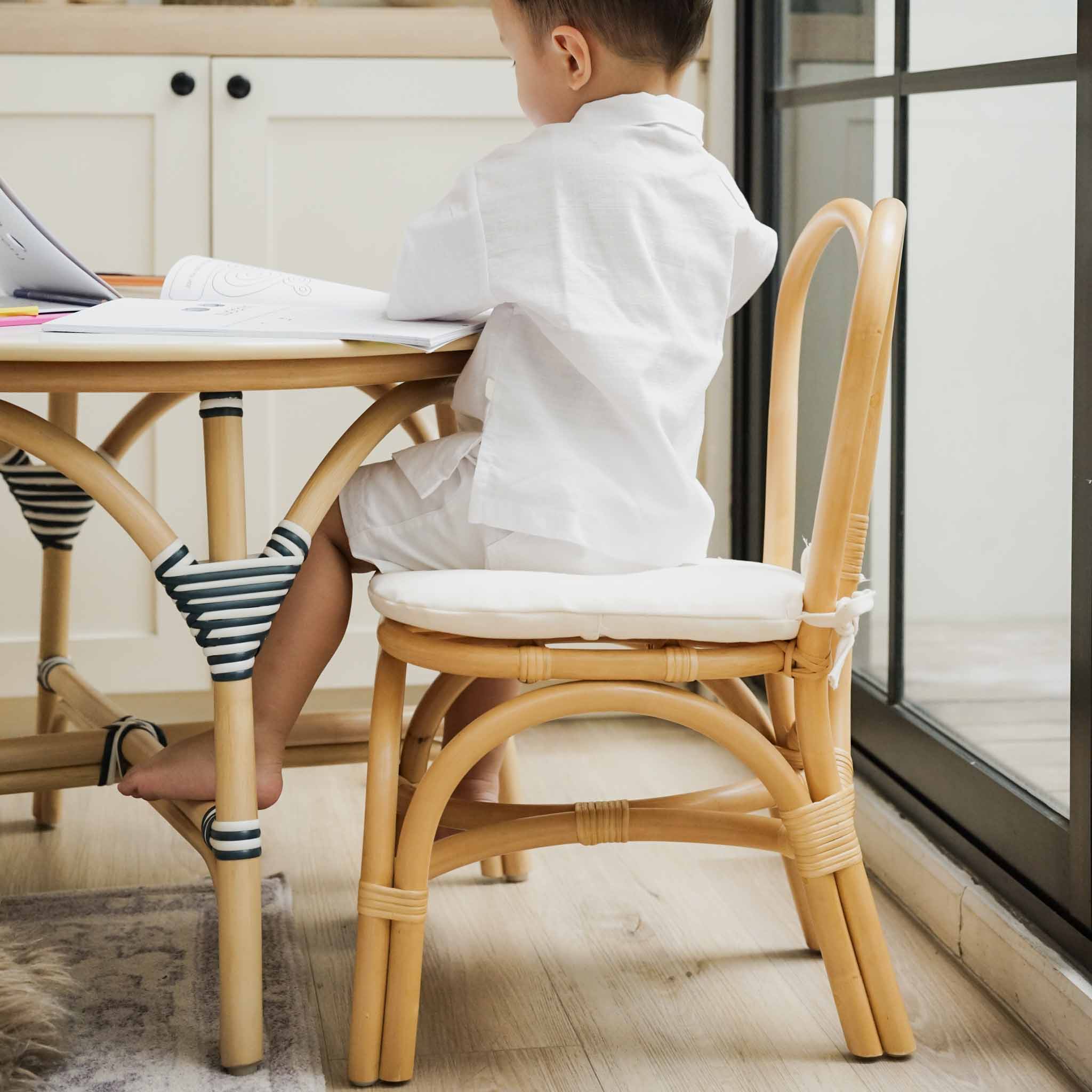 Child sitting on the natural handcrafted rattan wicker Bunny Chair by MOMIJI with comfortable cushion whilereading at a table.