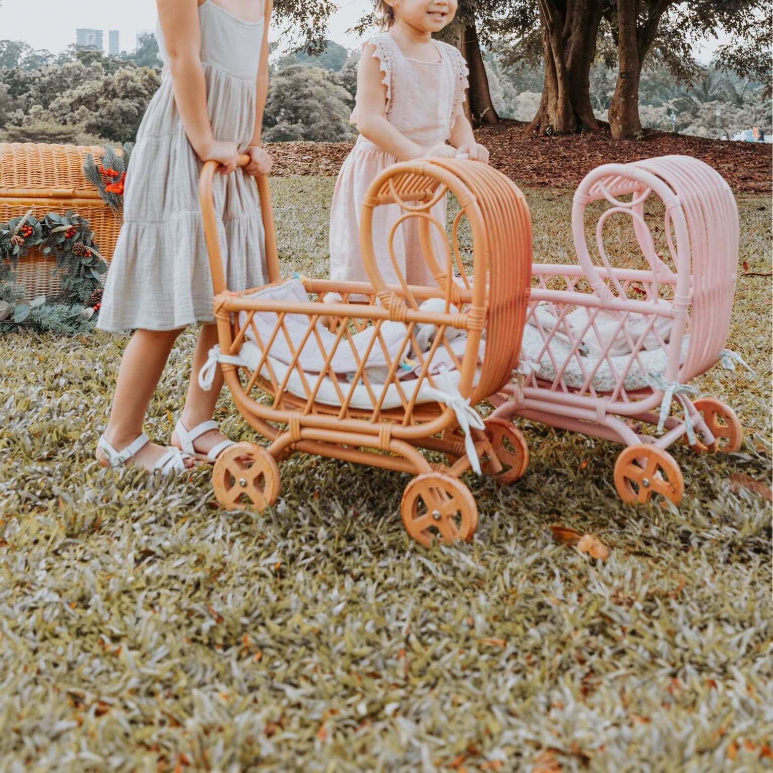 Two children pushing the handwoven rattan Layla Toy Stroller by MOMIJI in pink and natural versions while playing in a park.