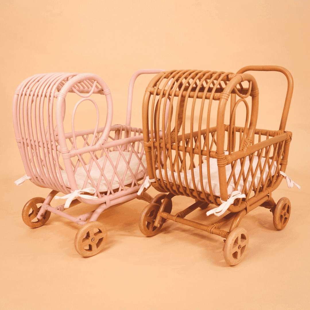 Handwoven rattan Layla Toy Stroller by MOMIJI in pink and natural versions with white seat pad inside, displayed side by side.