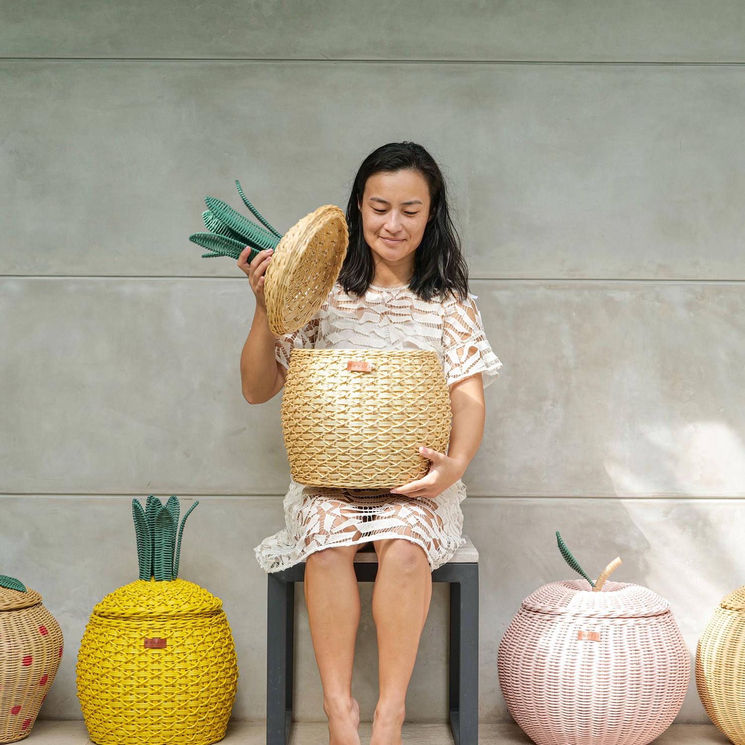 Woman Opening Pineapple Rattan Storage Basket by MOMIJI with fruit rattan basket collection on the floor.