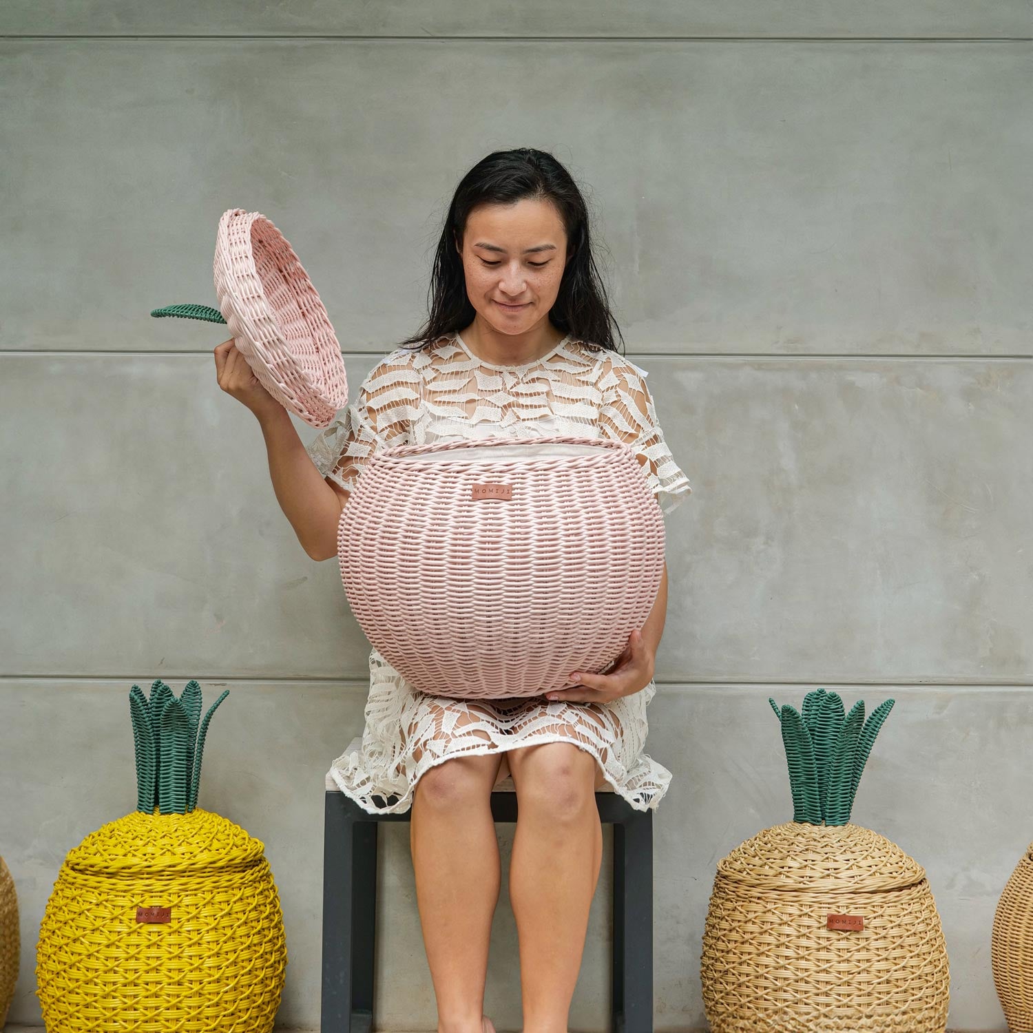 Woman opening a pink apple rattan storage basket by MOMIJI with both Yellow and Natural Pineapple rattan storage baskets on the floor.