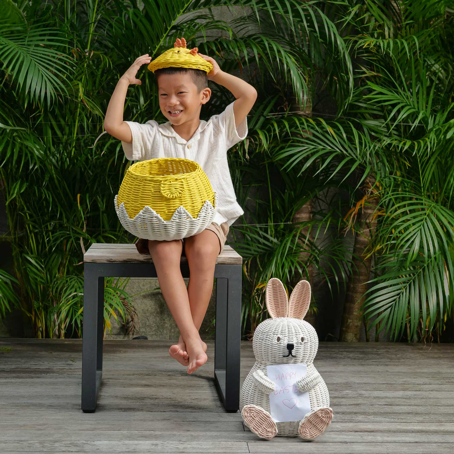 Child sitting on a stool with a Chick Rattan Storage Basket and Bunny Rattan Basket on the Floor by MOMIJI, surrounded by greenery.
