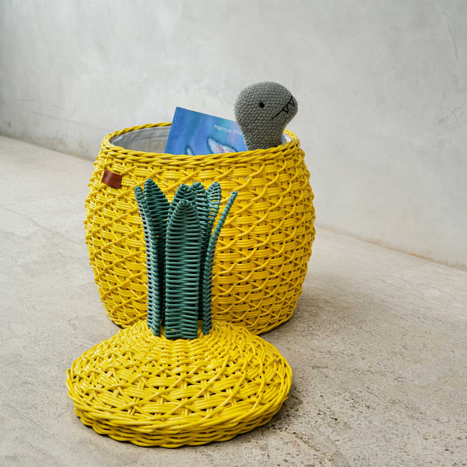 Side view of an opened yellow Pineapple Rattan Storage Basket with a book and a toy inside the basket, lid on the floor.