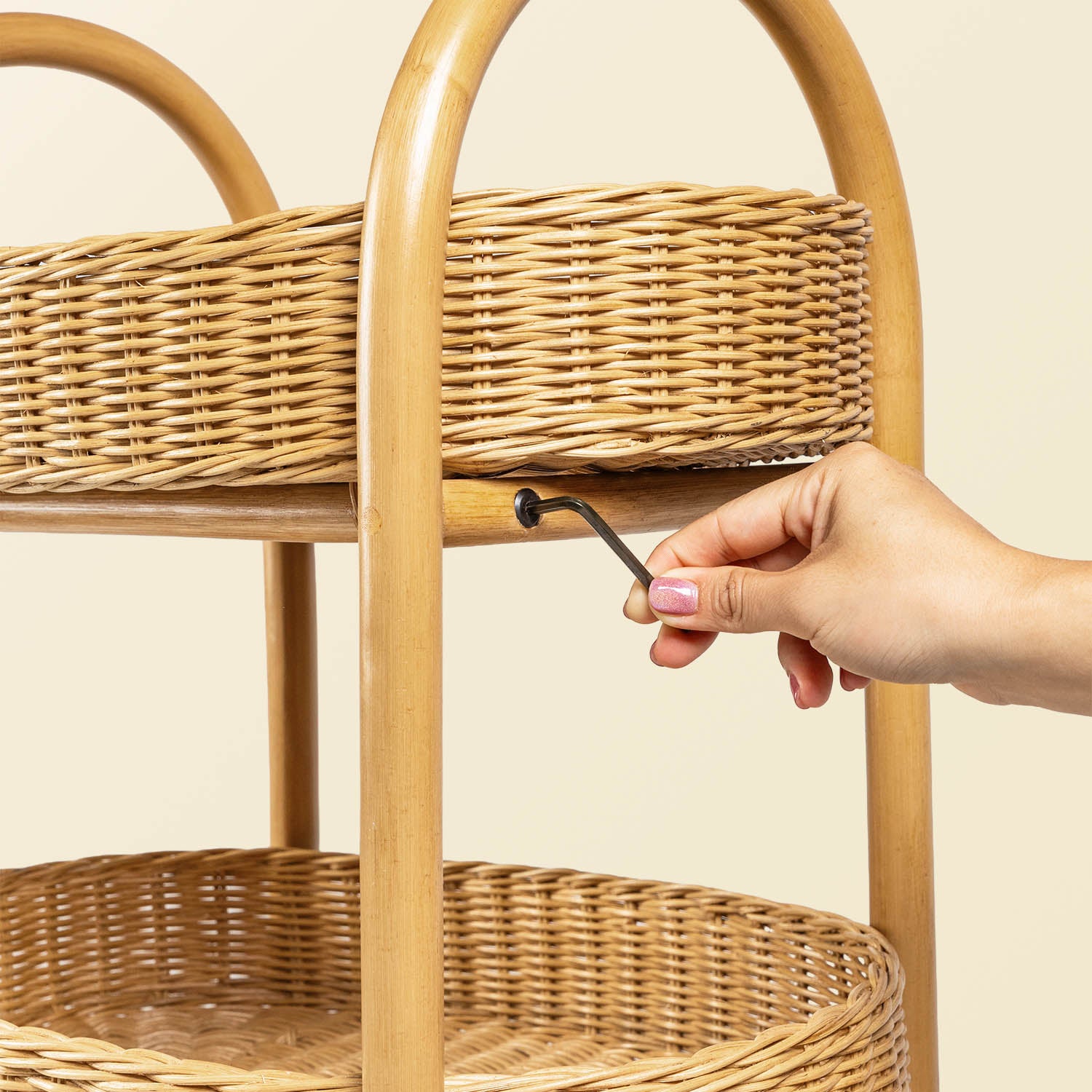 Hand assembling the utility cart with a hex key. Close-up of woven wicker trays and polished rattan pole structure frame with a neutral background
