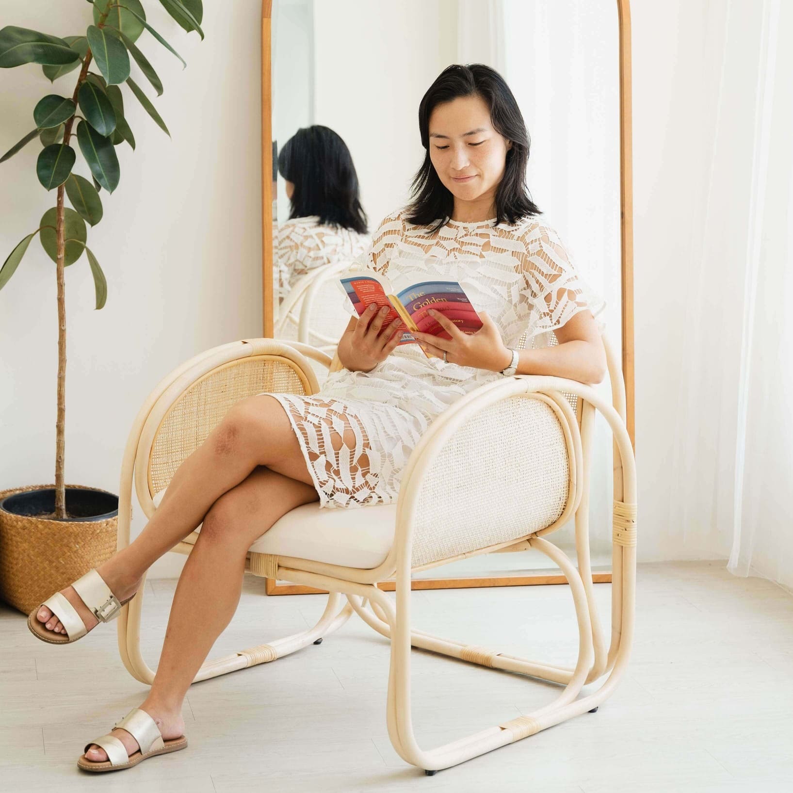 Woman relaxing on a handcrafted wicker Willow Lounge Chair by MOMIJI while reading a book in a bright room with indoor plants and a mirror.
