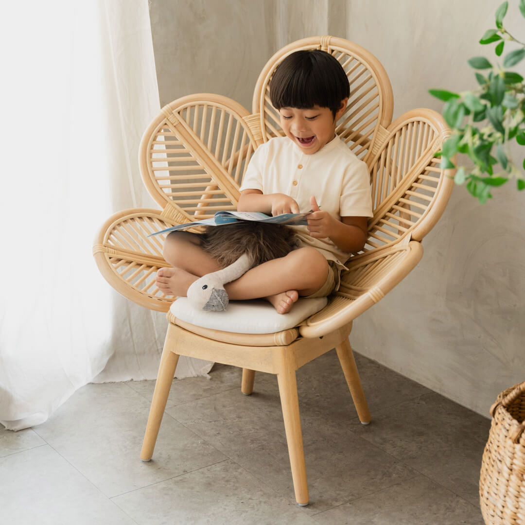 Smiling child sitting on the handcrafted rattan Petal Kids Chair by MOMIJI while reading a book indoors.
