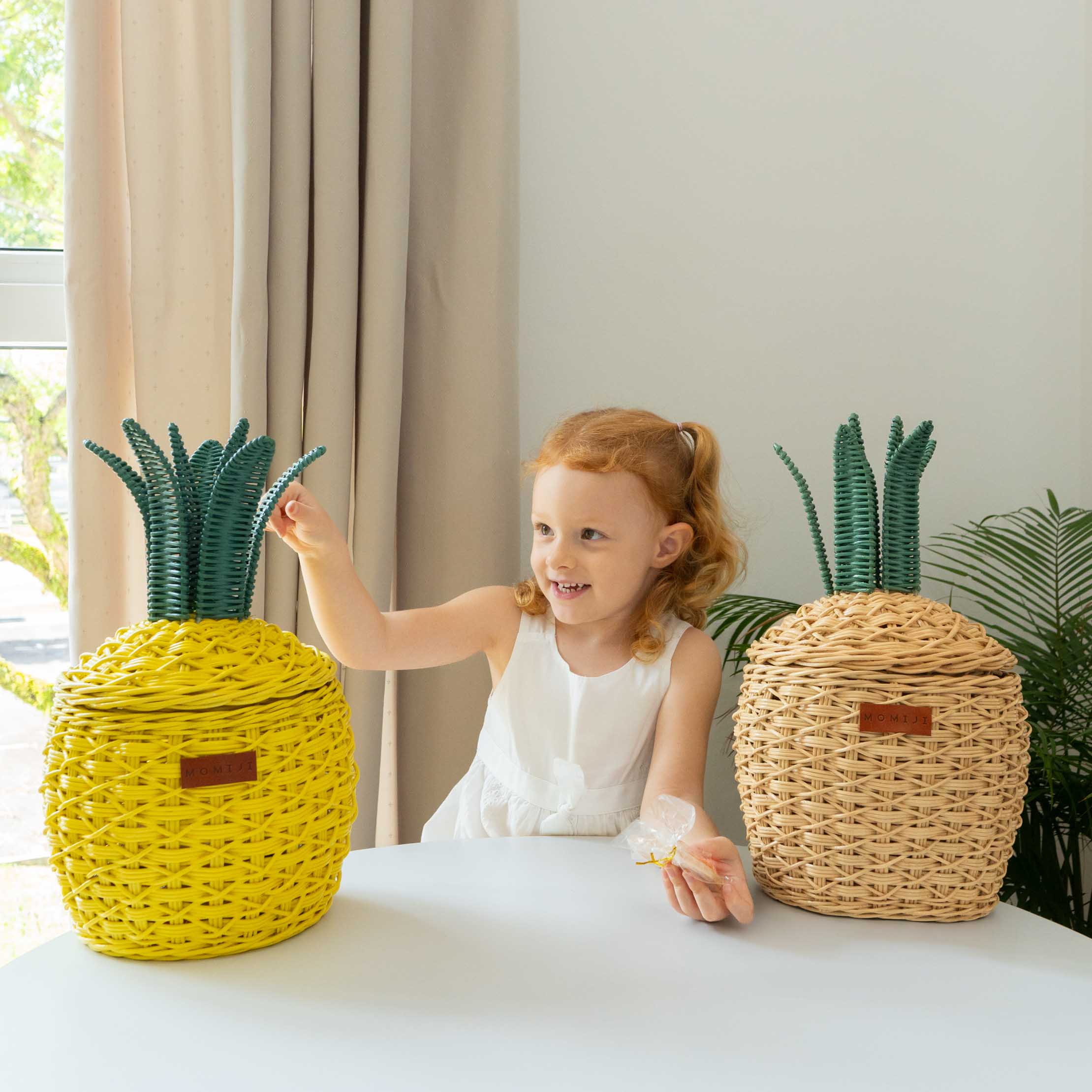 Child playing with Pineapple Rattan Storage Basket baskets on a table next to a natural colored Pineapple Rattan Storage Basket.