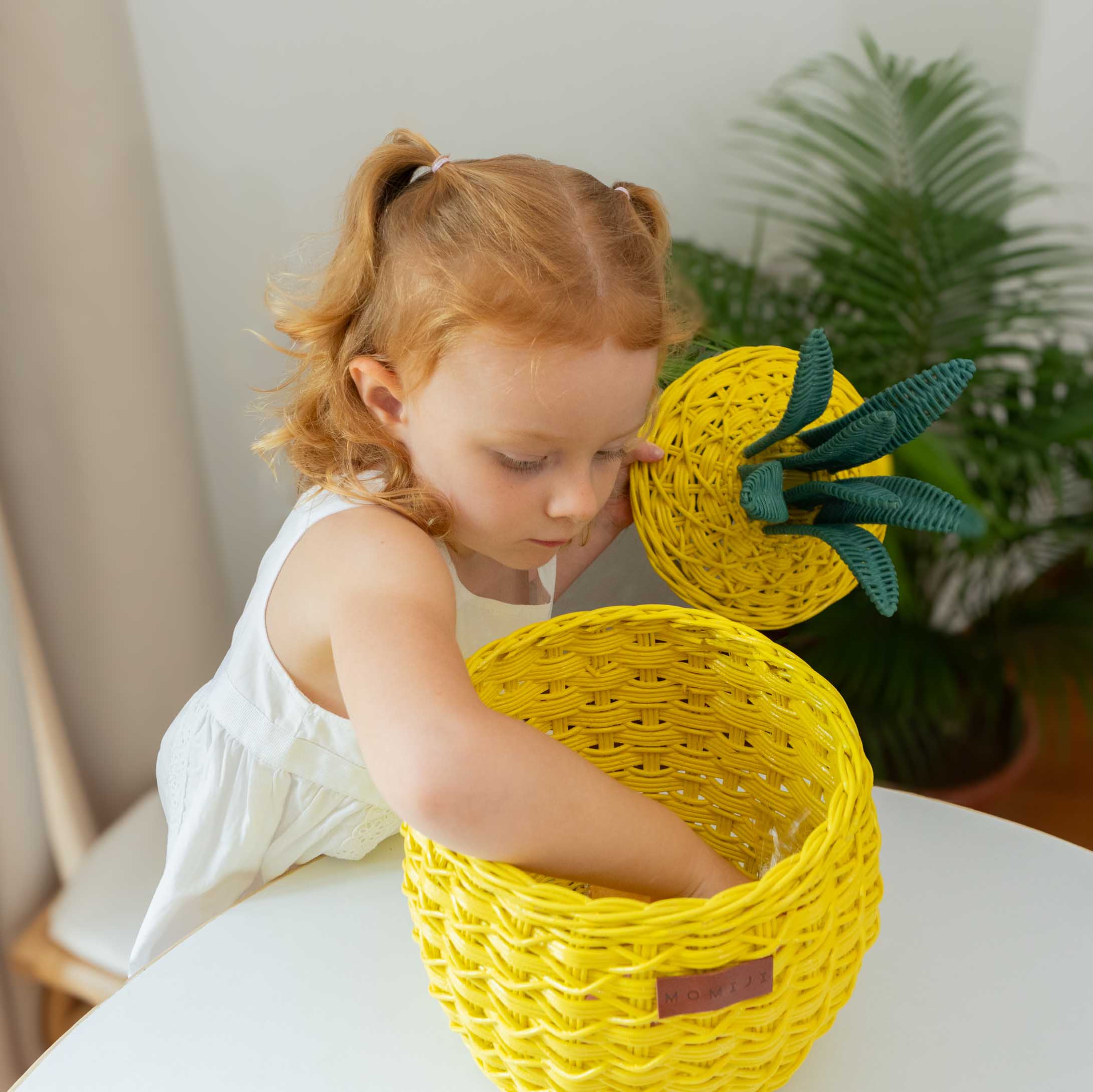 Child open Pa handcrafted yellow pineapple rattan storage basket in small size by MOMIJI in a bright room with a plant in the corner.