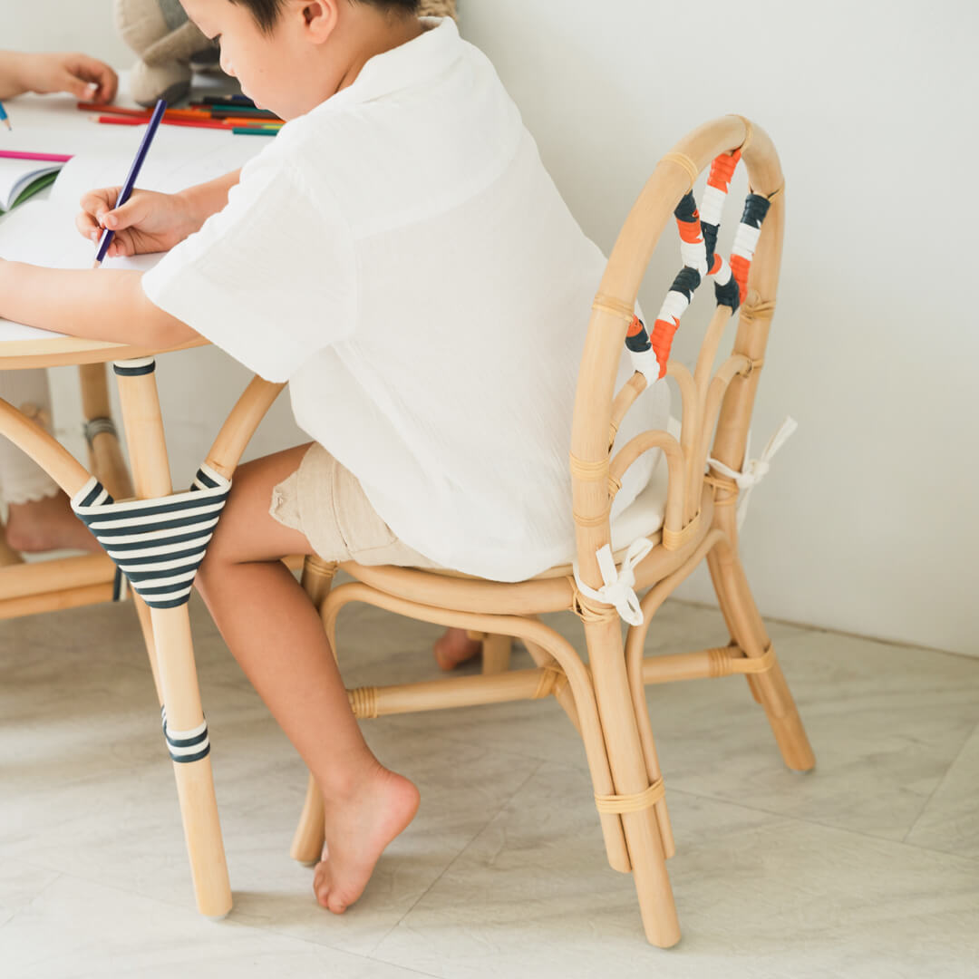 Child sitting on a handcrafted rattan wicker Quentin Kids Chair by MOMIJI while writing at a small table, supporting comfortable kids activities.