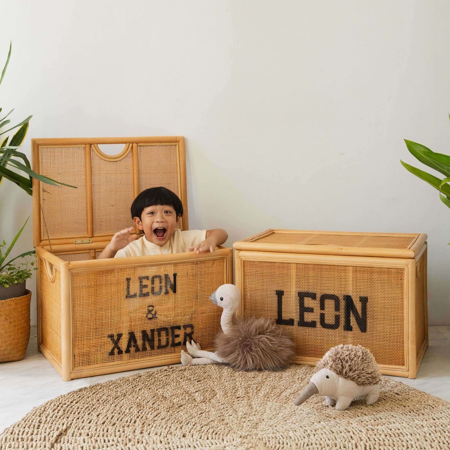 Child playing with wicker storage trunks labeled “LEON & XANDER” by MOMIJI in a room with indoor plants, with an extra trunk beside.