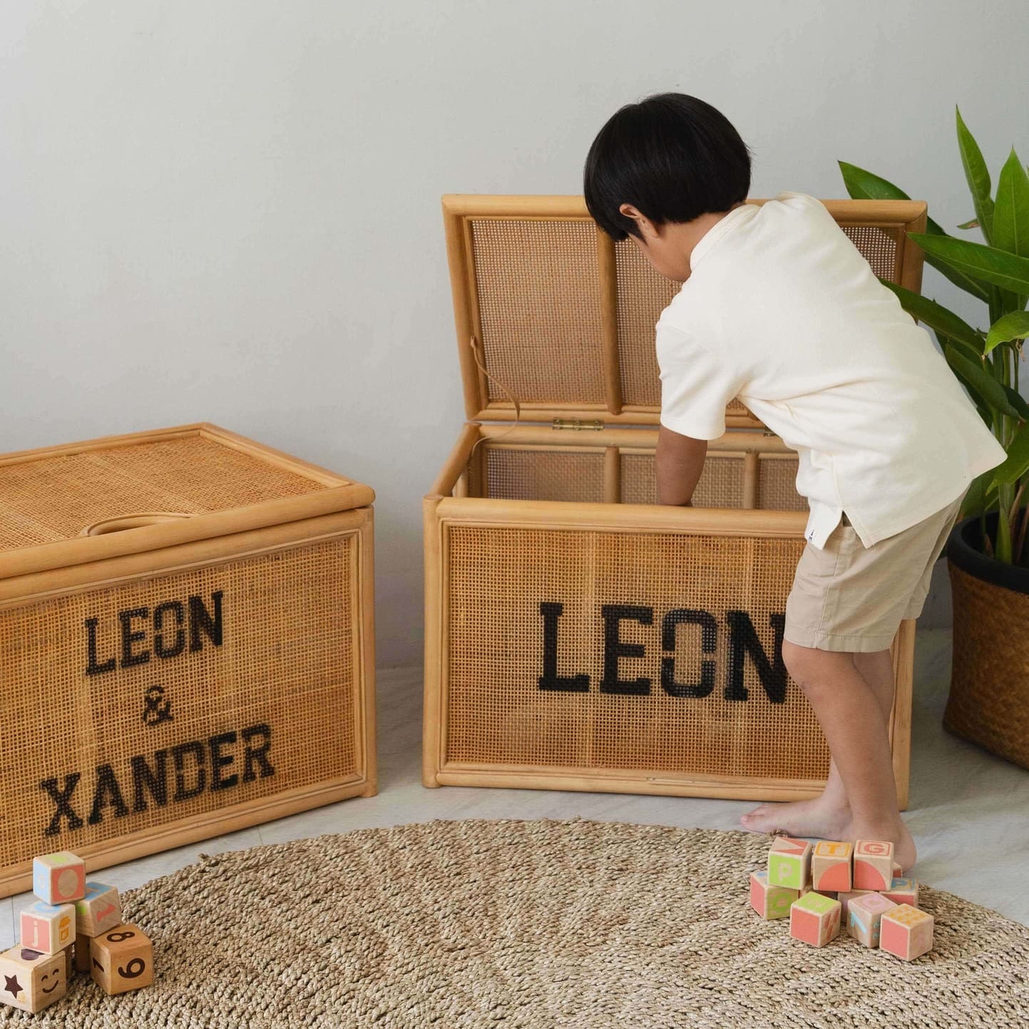 Child playing with colorful blocks next to wicker storage trunks labeled “Leon & Xander” by MOMIJI on a light gray floor.