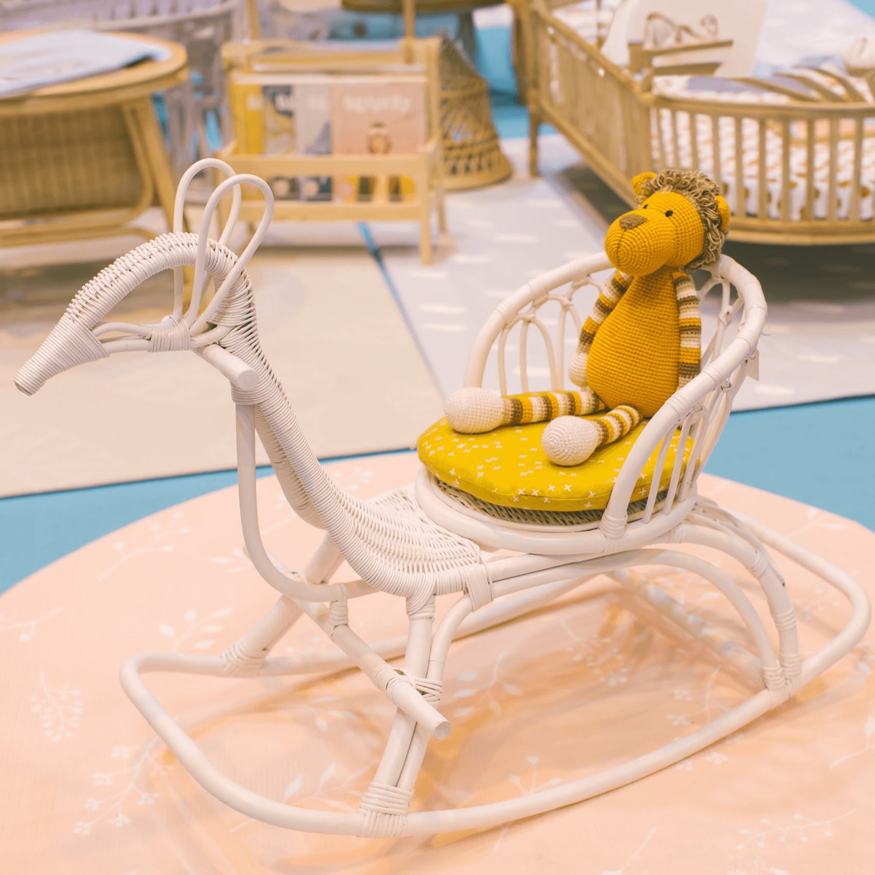 Doll sitting on the Rocking Peacock by MOMIJI in natural color with a yellow cushion, placed on a rug in a room filled with rattan furniture.