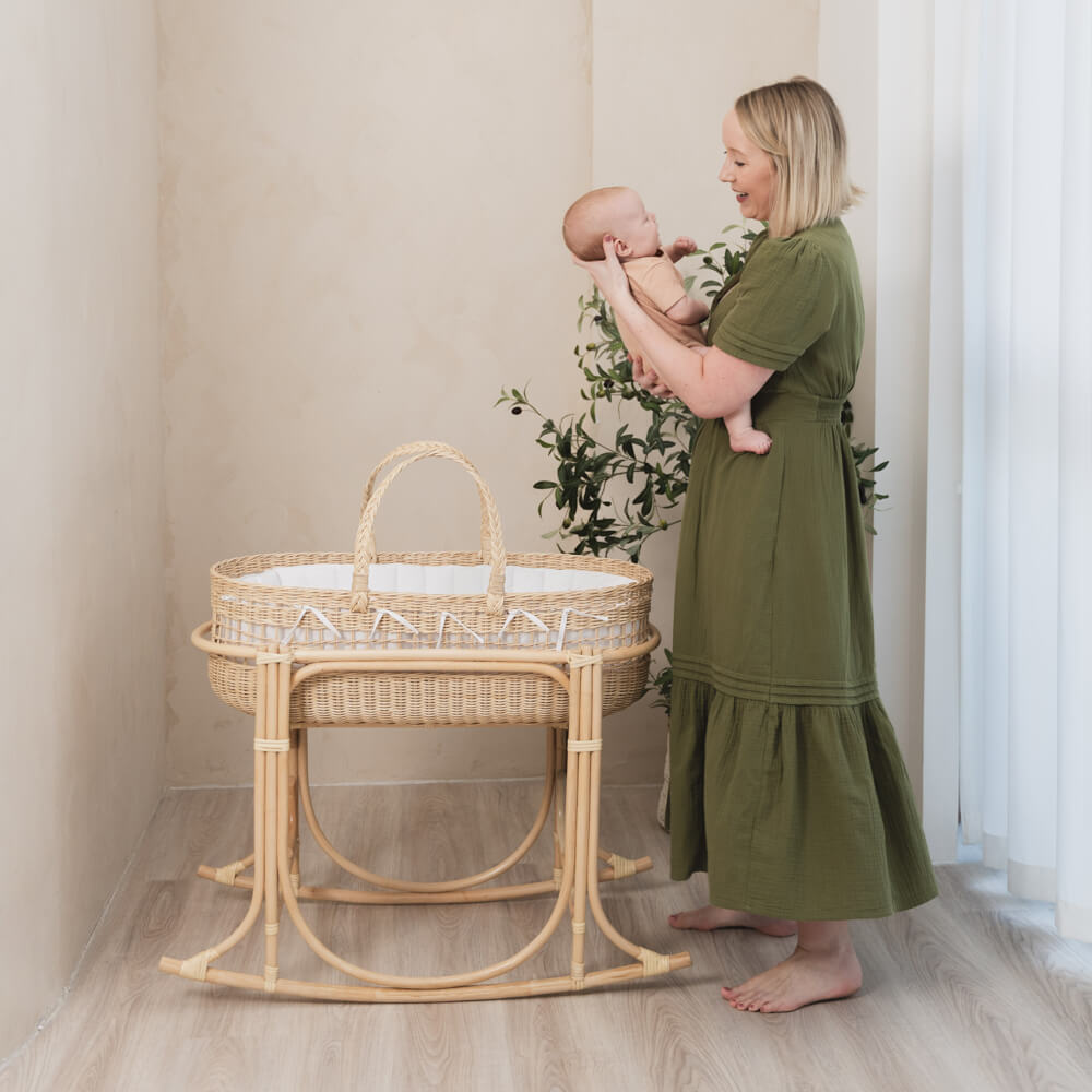 Woman holding a baby next to a wicker Moses basket on a rocking stand by MOMIJI in a softly lit room.
