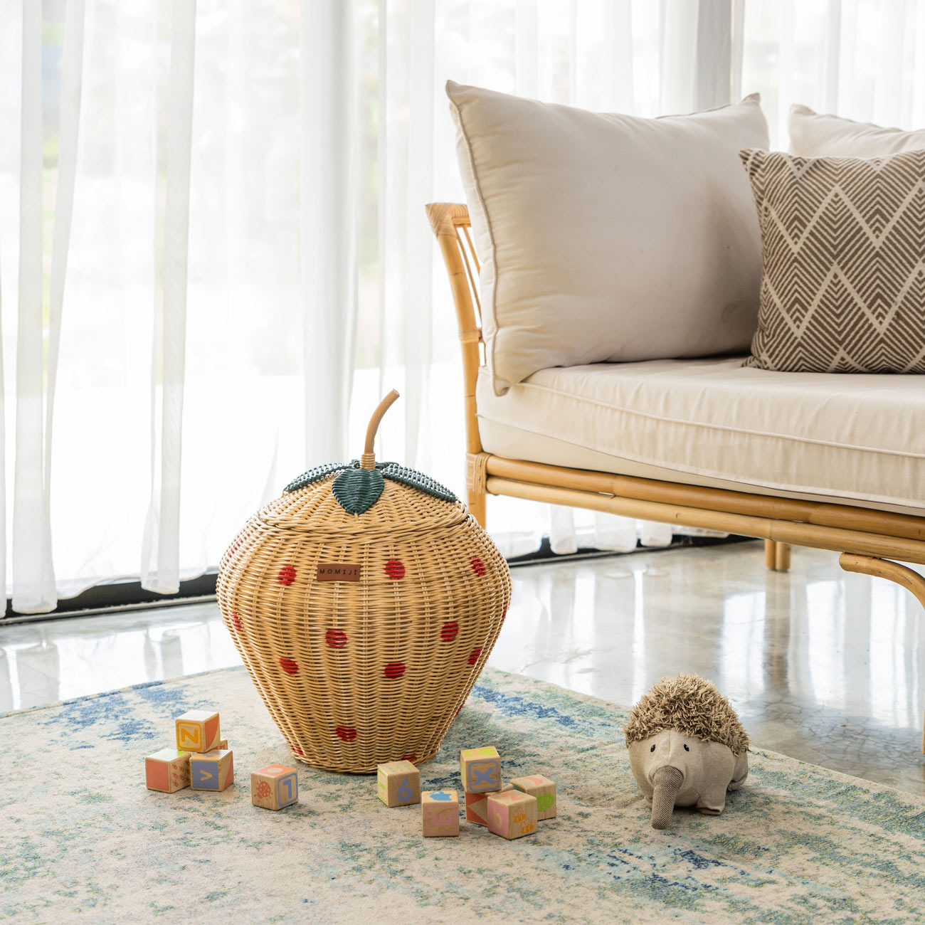 Handcrafted strawberry rattan wicker pineapple storage basket by MOMIJI, featuring a green leaf and natural stem lid, styled in a living room with children’s toys and a rattan bench.