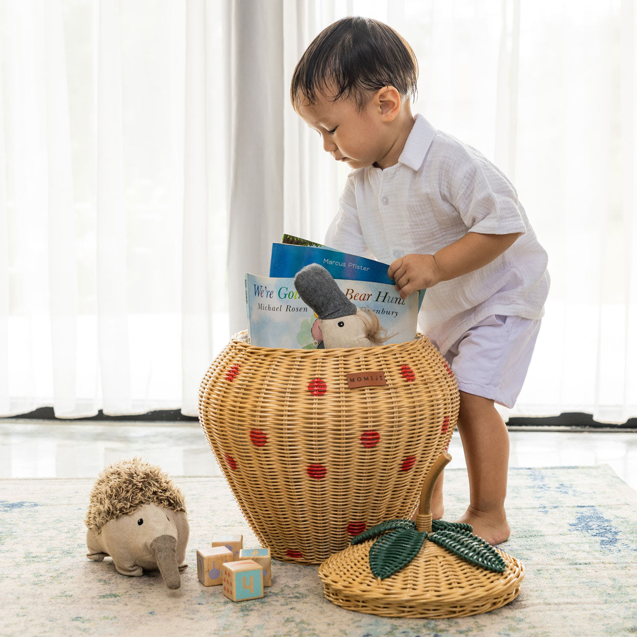 Child placing a book into the Strawberry Rattan Storage Basket by MOMIJI with toys around, shown with the lid removed in a bright kids room.