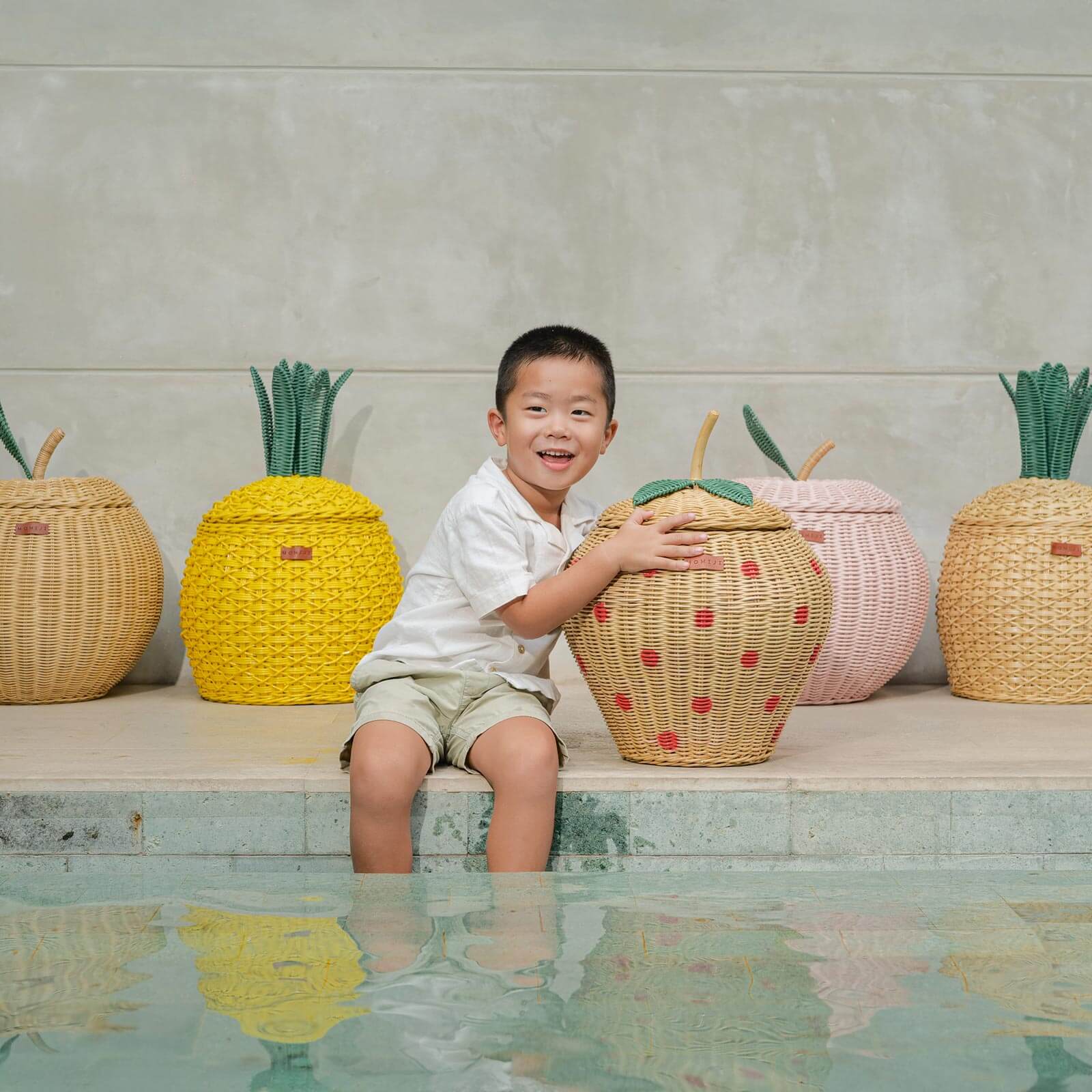 Child sitting by a pool holding a Strawberry Rattan Storage Basket with fruit designed rattan storage basket collection in the background.