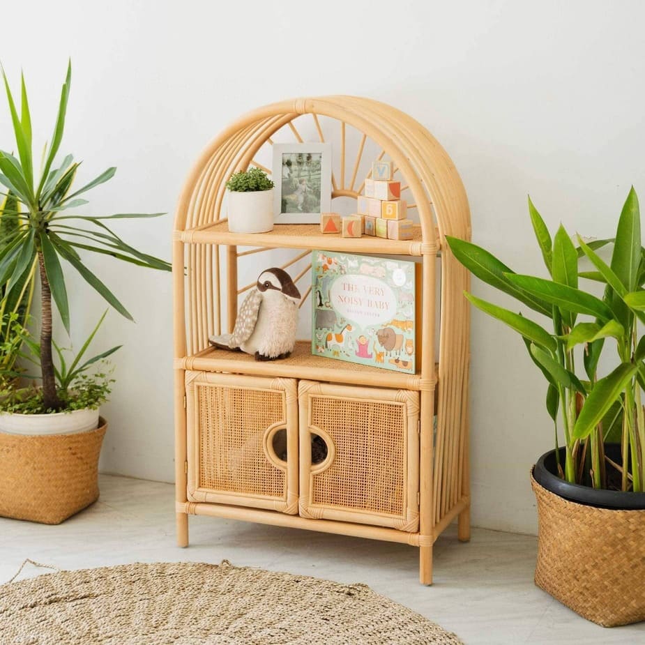 Angled view of the handcrafted rattan Sunshine Arch Cabinet Shelf by MOMIJI filled with children’s toys in a room with multiple potted plants.