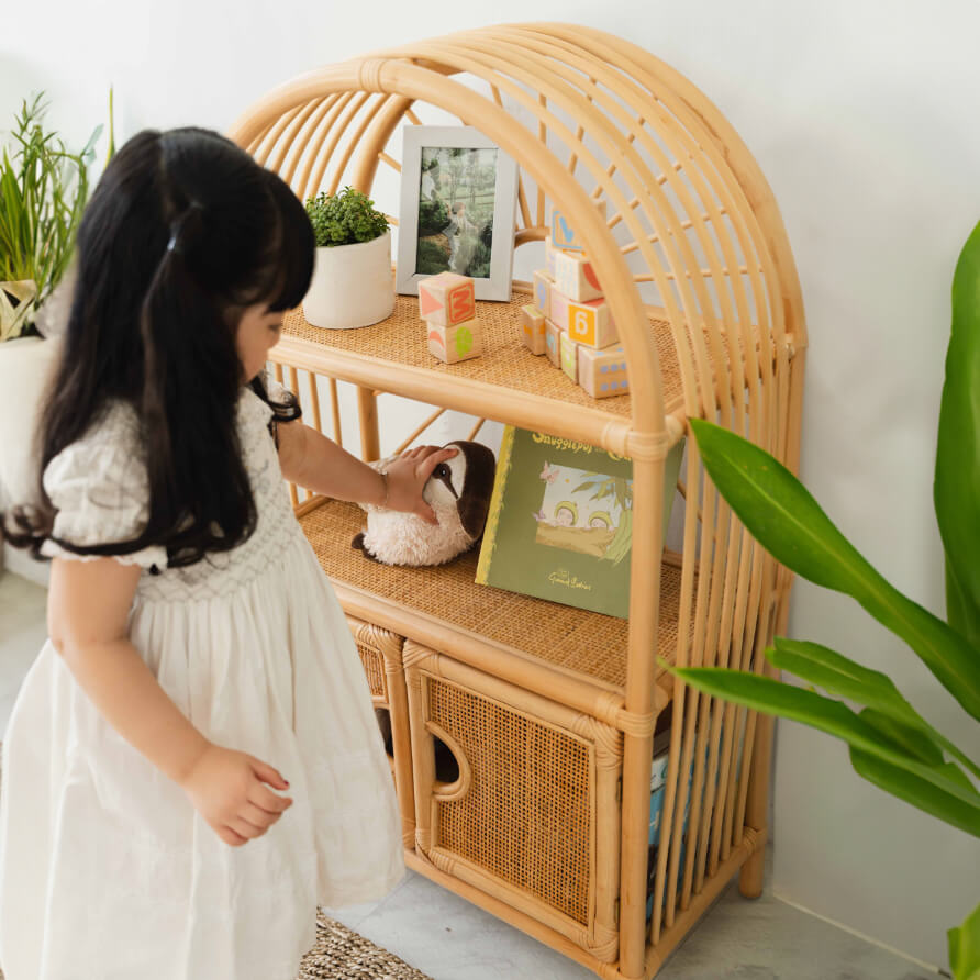 Child taking a doll from the handcrafted rattan wicker Sunshine Arch Cabinet Shelf by MOMIJI in a room with plants.