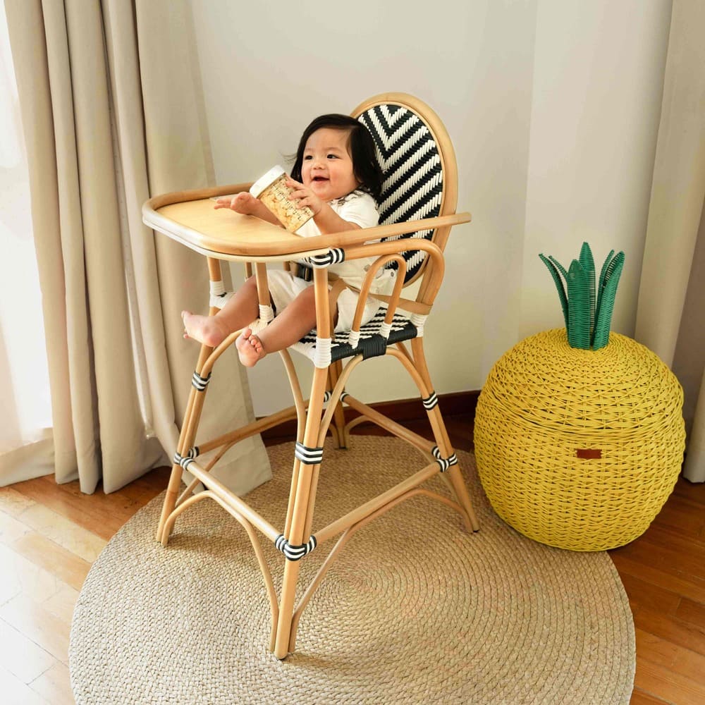 Laughing child holding a tumbler while sitting on the Handcrafted rattan wicker Tumbuh Kids High Chair by MOMIJI indoors.