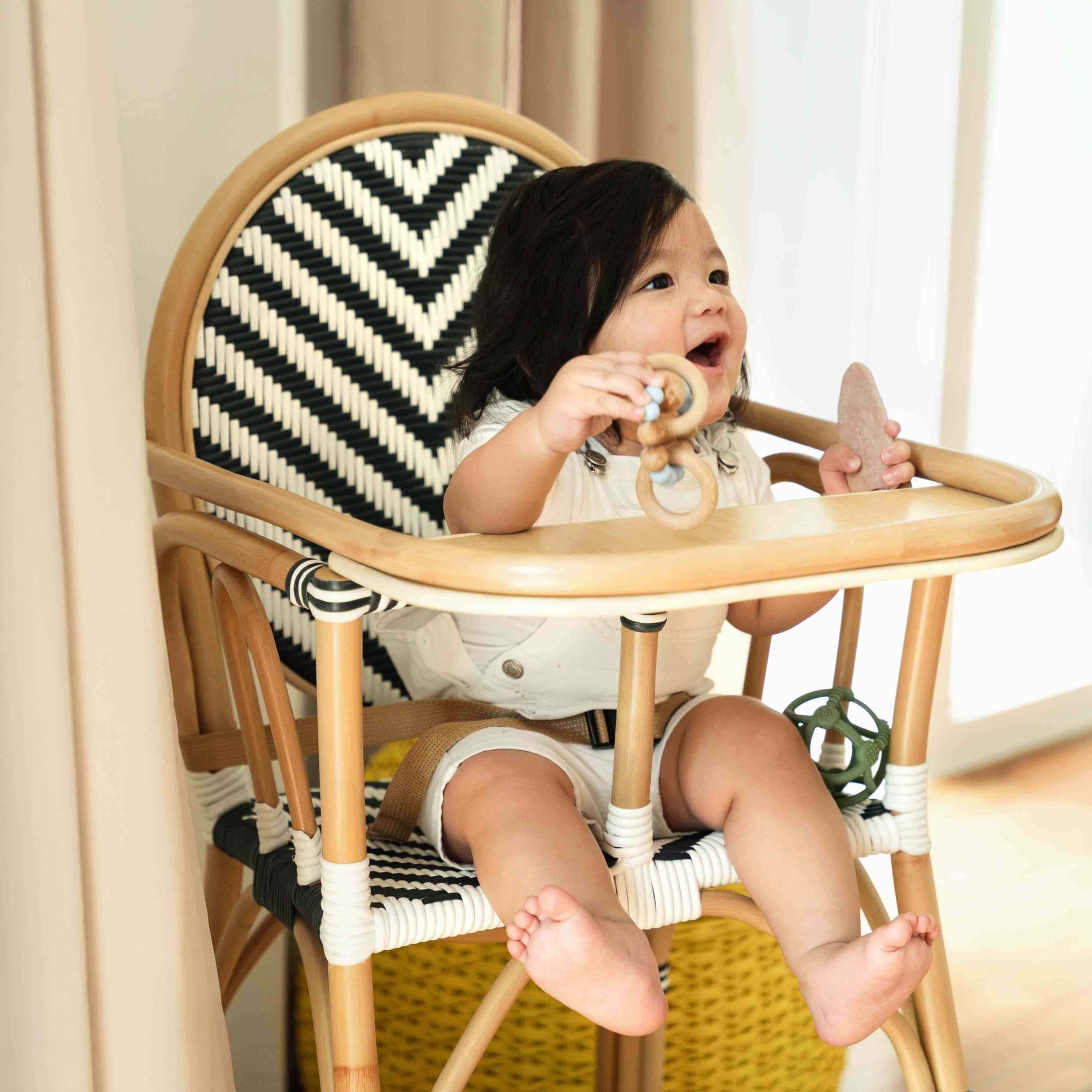 Smiling child sitting on the Handcrafted rattan wicker Tumbuh Kids High Chair by MOMIJI holding snacks and toys.