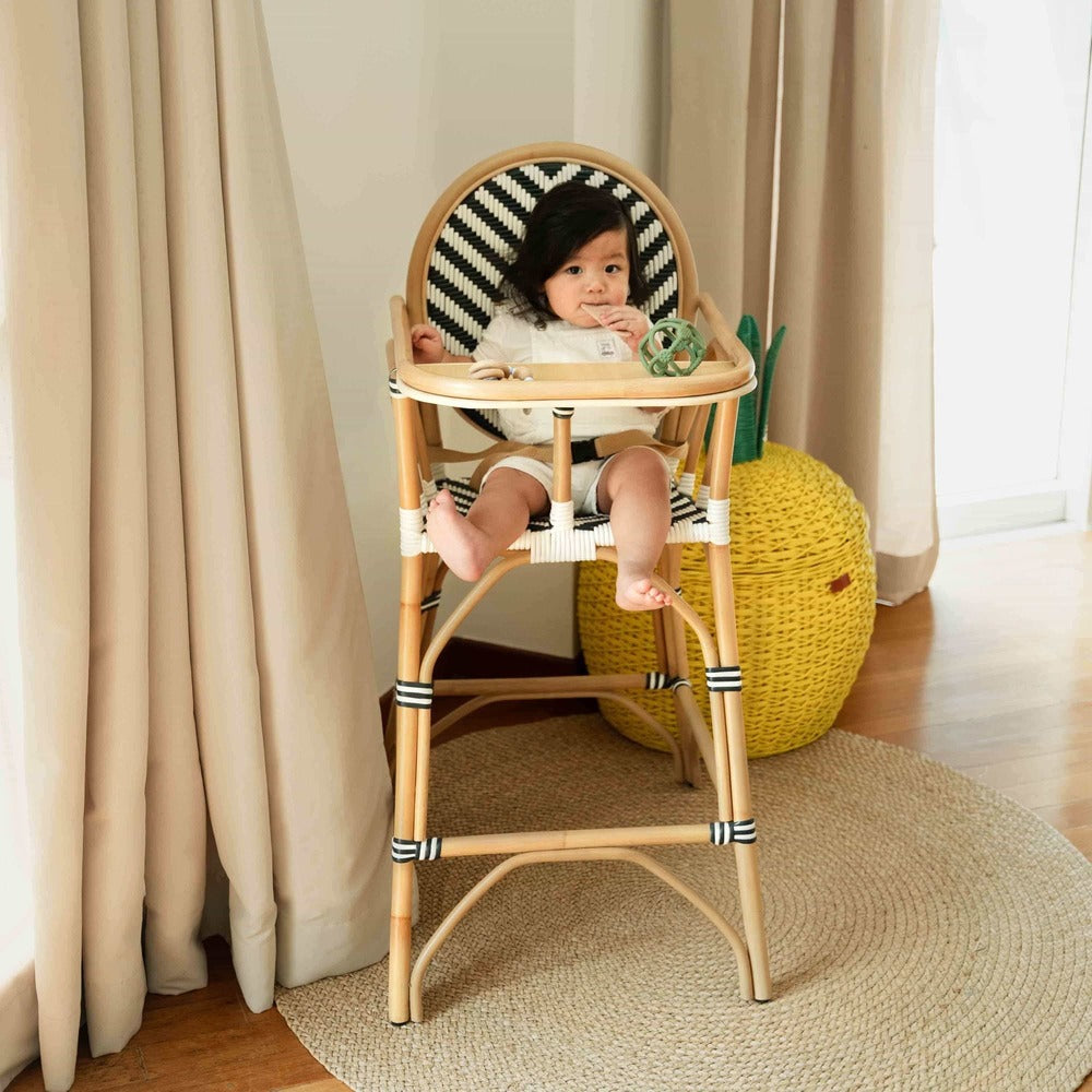 Child sitting on the Handcrafted rattan wicker Tumbuh Kids High Chair by MOMIJI eating snacks in a room with a pineapple basket.