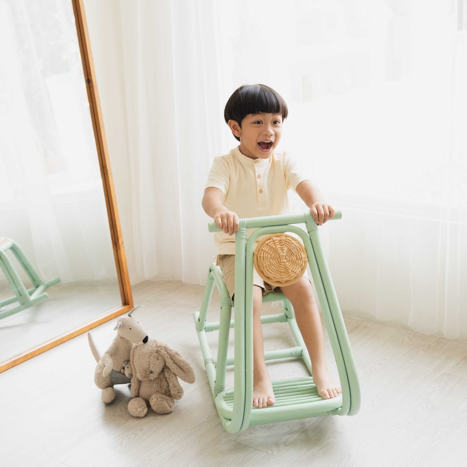 Happy child riding the Vespa Rocker – Mint by MOMIJI on the floor with toys in a playroom, showcasing the mint green frame and woven rattan seat.