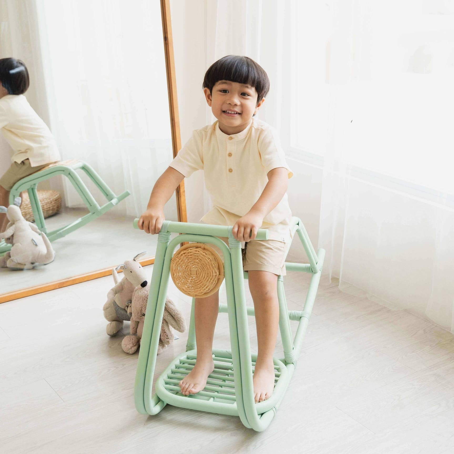 Child playing with a green vespa rocking chair in a room with a mirror and stuffed animals.