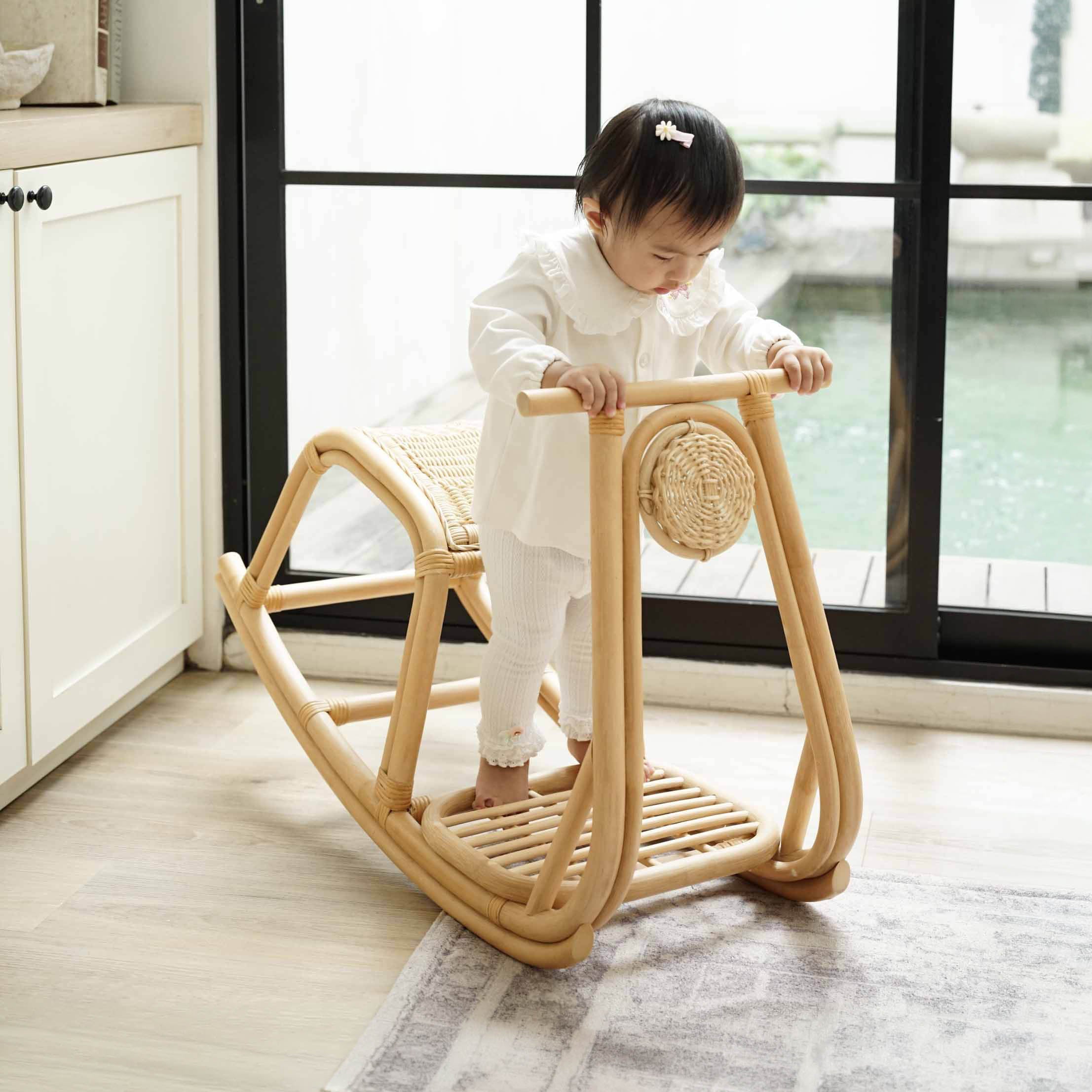 A toddler playing on the handcrafted natural rattan Vespa Rocker by MOMIJI in a bright indoor room with large black-framed windows and light wood flooring, encouraging imaginative and active play.