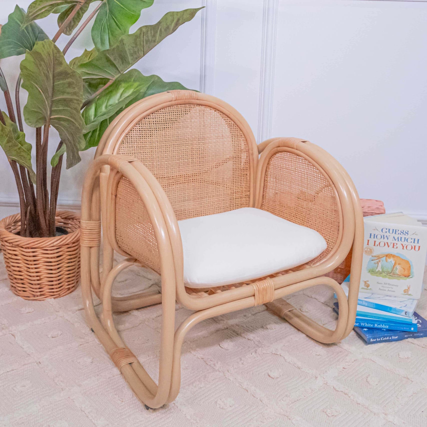 Child-sized rattan Willow Lounge Chair by MOMIJI with a white cushion next to books and a plant on a wooden floor.