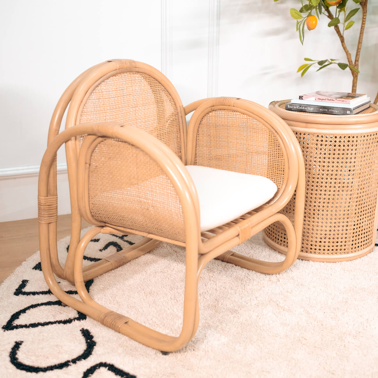 Child-sized rattan Willow Lounge Chair by MOMIJI with a white cushion in a room with a rug and small table, showcasing its sturdy structure and natural woven texture.